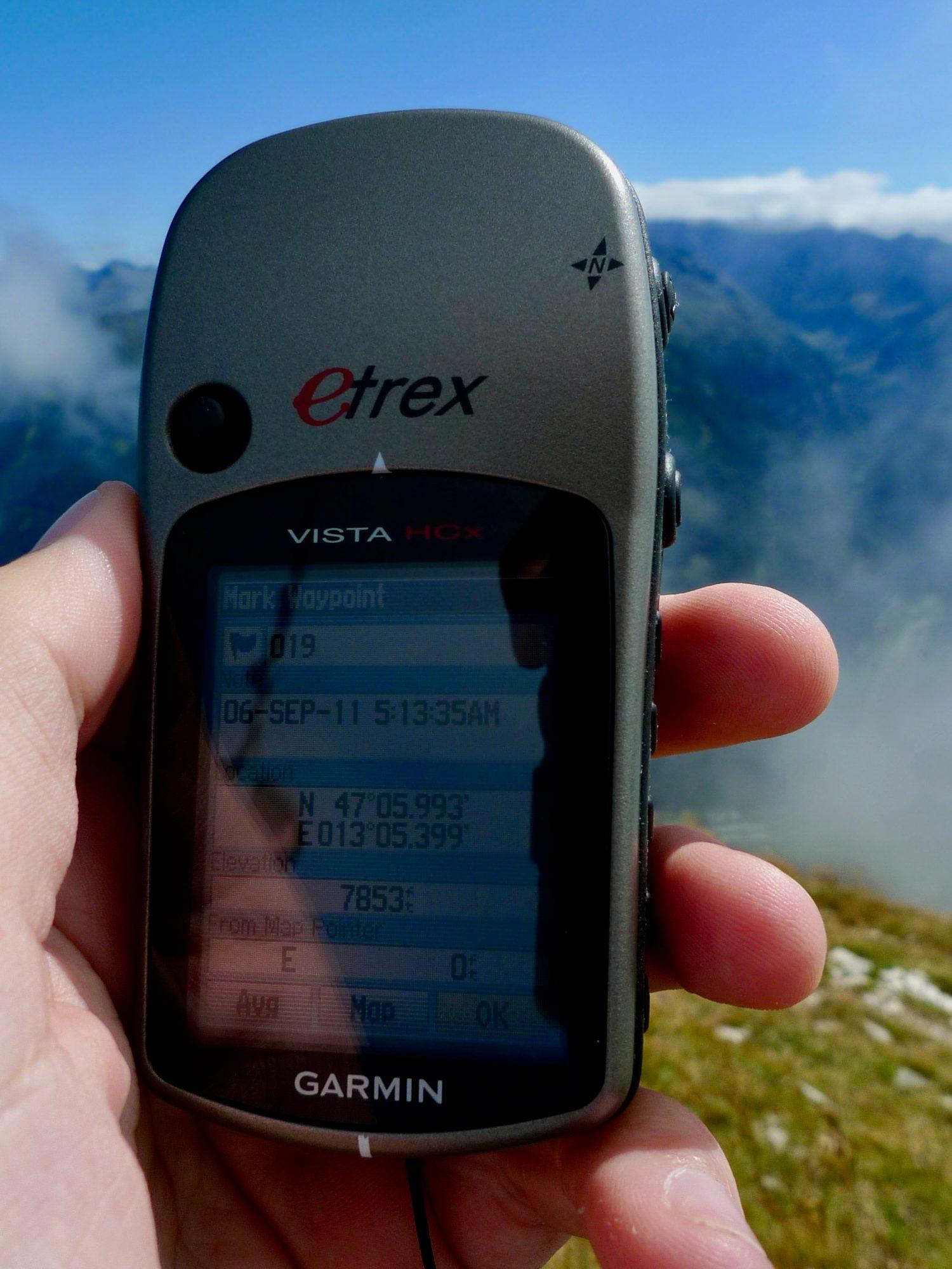 A Garmin eTrex Vista HCx handheld GPS held up in one hand at altitude, its small grayscale screen showing a marked waypoint with coordinates and elevation, hazy mountains behind