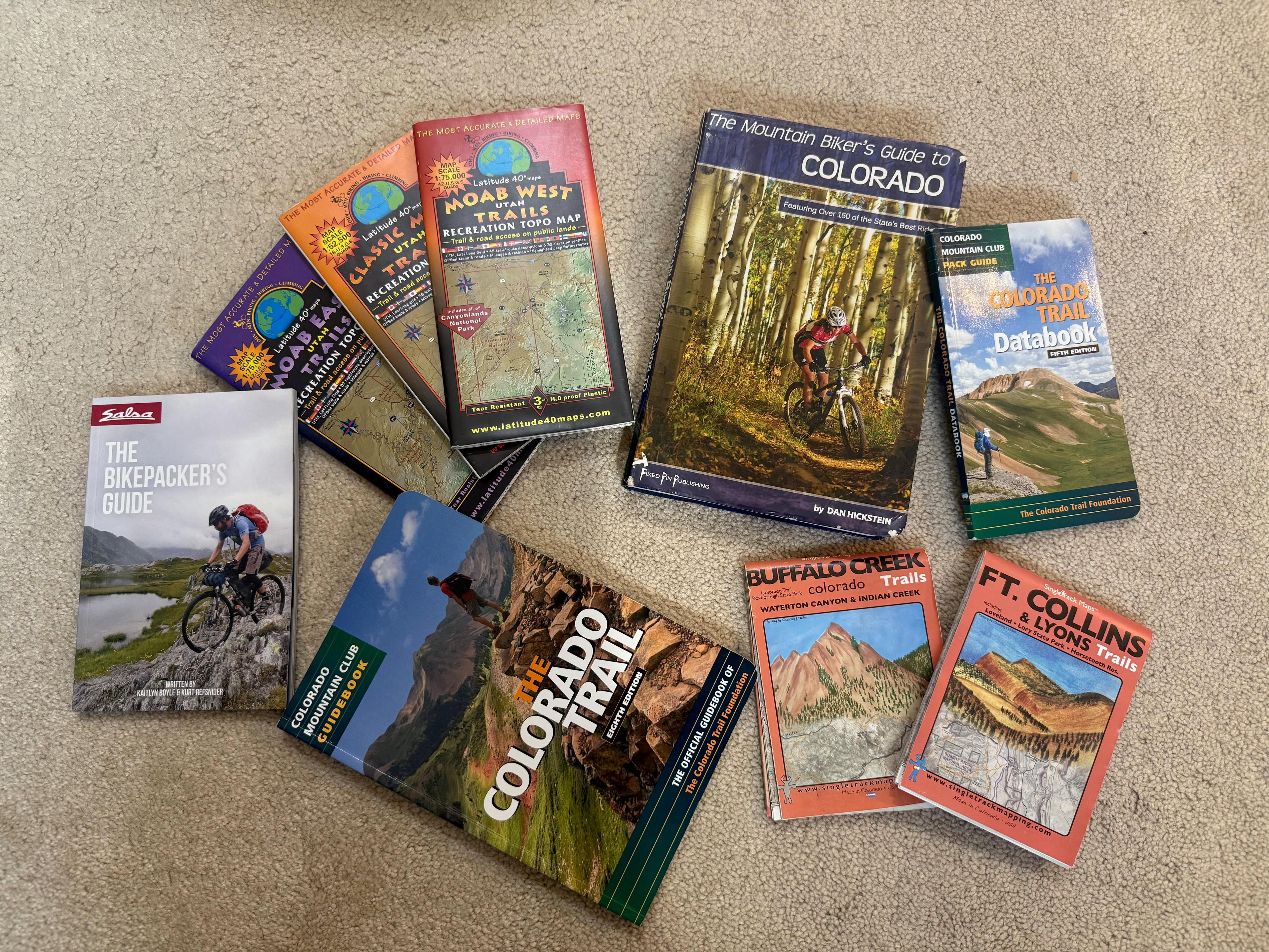 A fan of paper mountain-bike trail maps and guidebooks spread across carpet, including Latitude 40 Moab maps, The Mountain Biker's Guide to Colorado, the Colorado Trail Databook, and Buffalo Creek and Ft. Collins trail maps