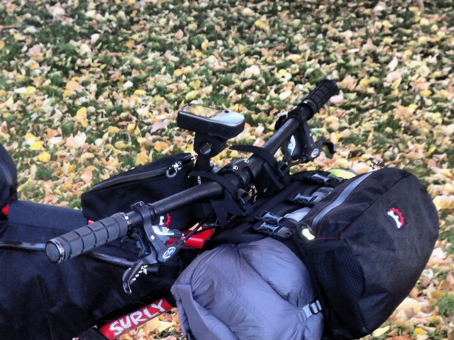 A red Surly bike lying on autumn leaves, loaded with a handlebar bag, a frame bag, and a sleeping bag strapped up front, with a small GPS computer mounted on the handlebar