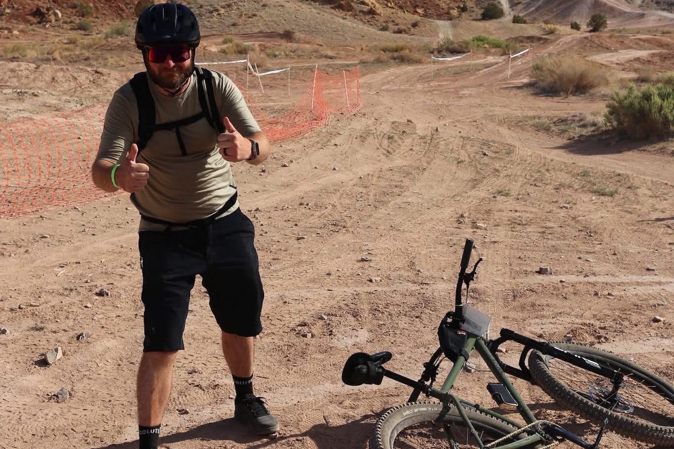 Rider giving a thumbs up next to a single-speed mountain bike laid over on a desert singletrack trail