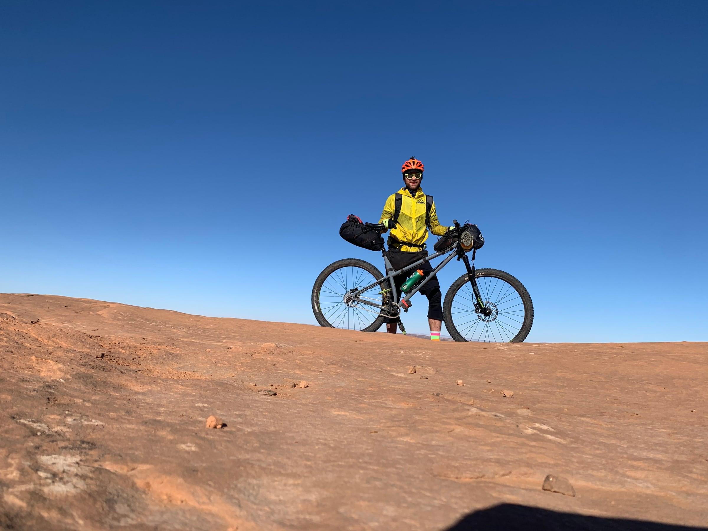 Rider standing with a loaded single-speed bikepacking mountain bike on slickrock, under a clear blue sky