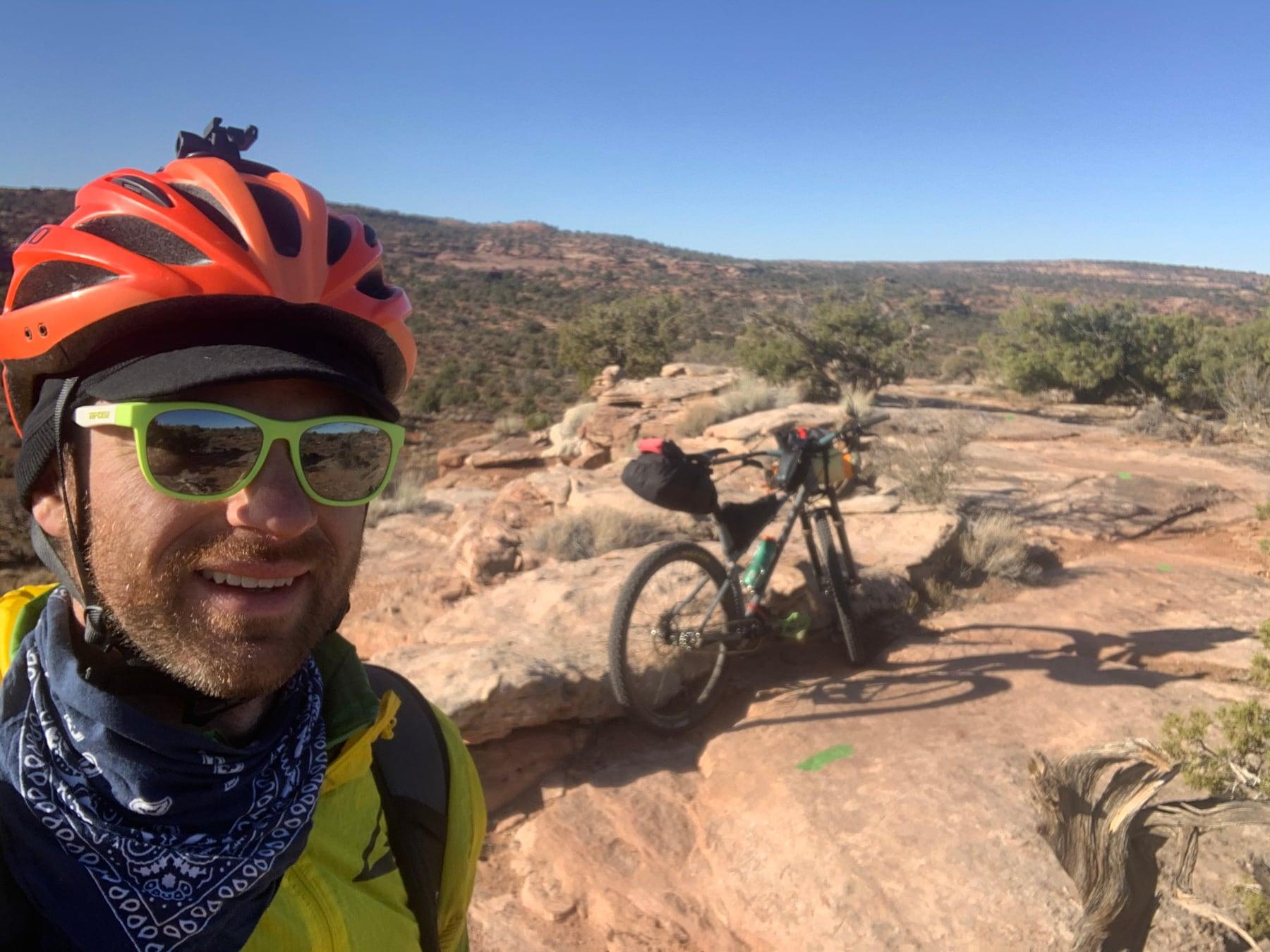 Close-up selfie of a rider in a helmet, sunglasses, and bandana, with a loaded bikepacking bike resting on slickrock behind him