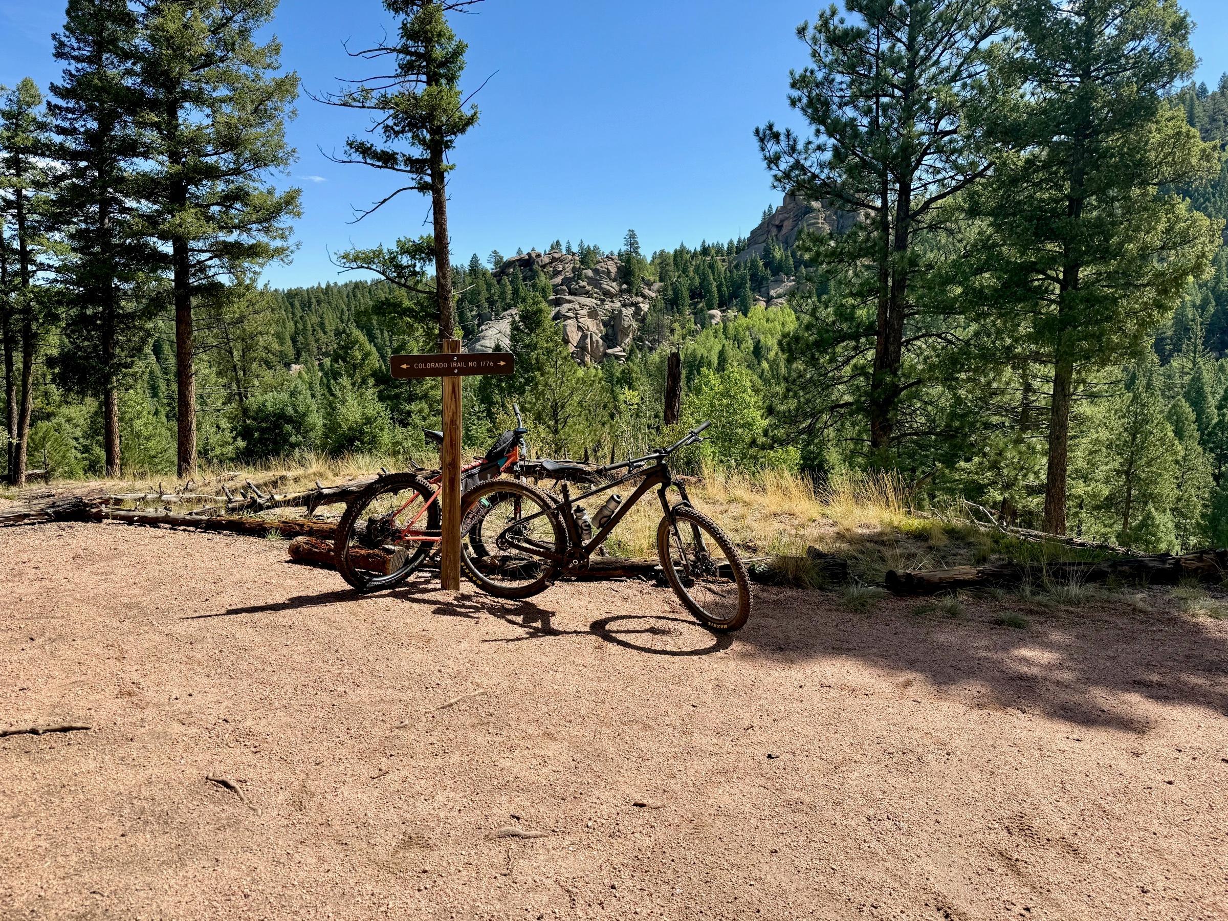 Two mountain bikes leaning against a wooden signpost marked Colorado Trail No. 1776, with a granite rock formation and pine forest in the background
