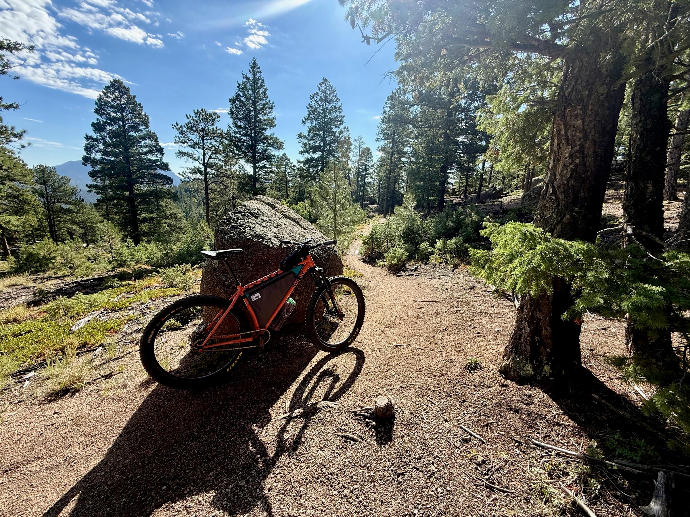 An orange single-speed Surly with a frame bag leaning against a granite boulder on a pine-forested singletrack trail, with distant mountains visible through the trees