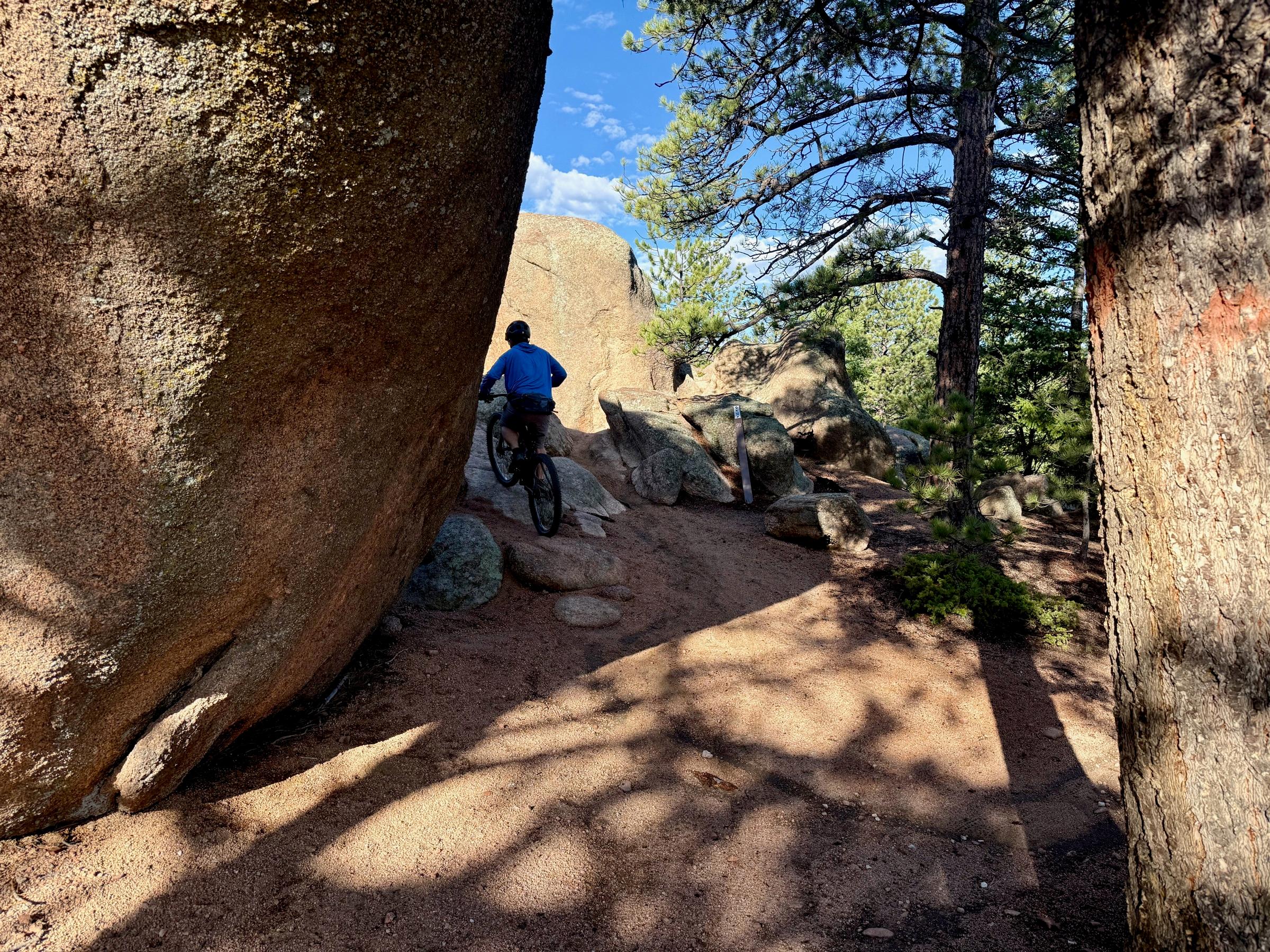 A cyclist in a blue jacket riding through a narrow rock passage between two large granite boulders on a singletrack trail