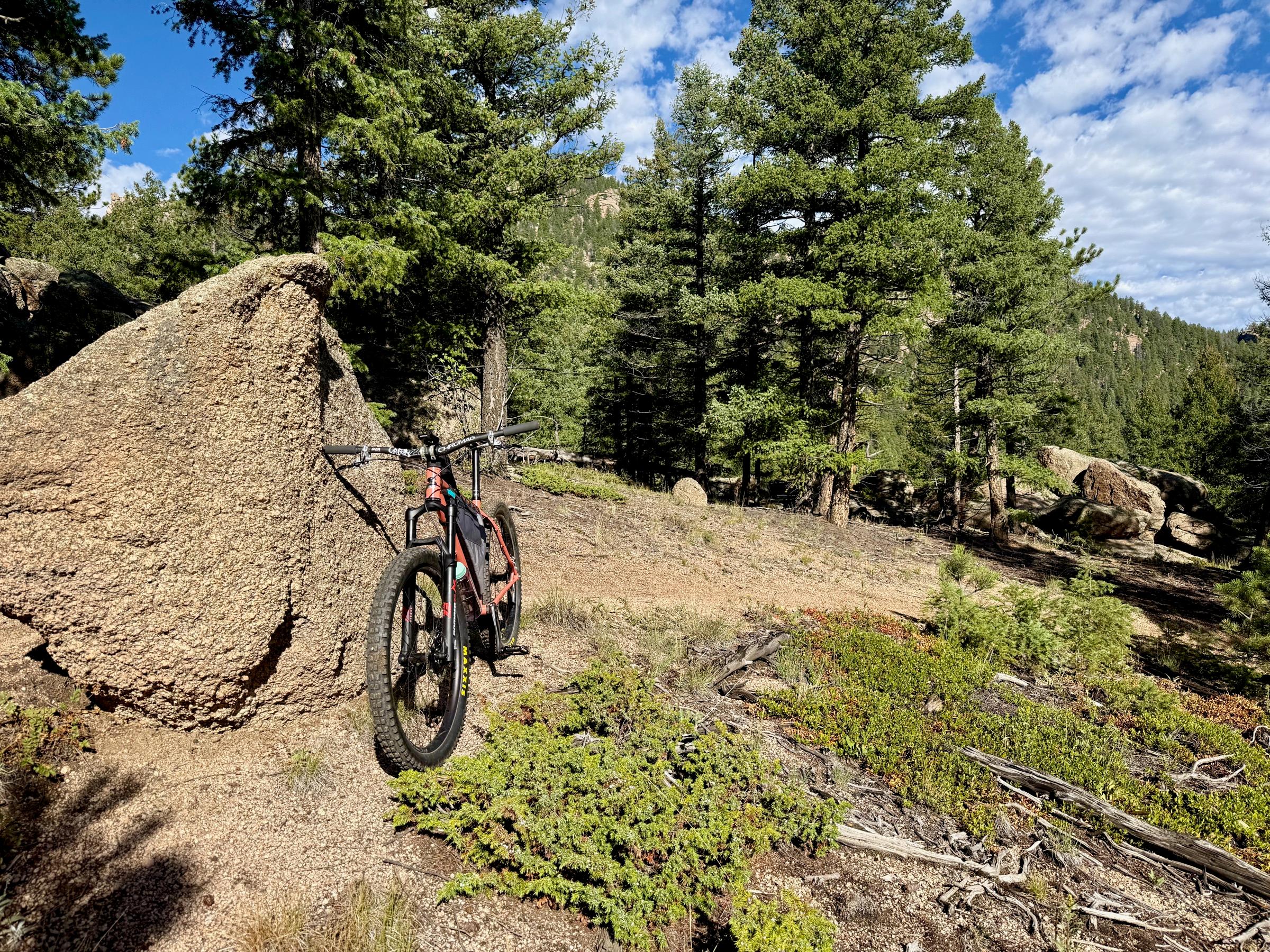 An orange single-speed mountain bike with a frame bag leaning against a granite rock slab, with pine forest and a rocky hillside in the background