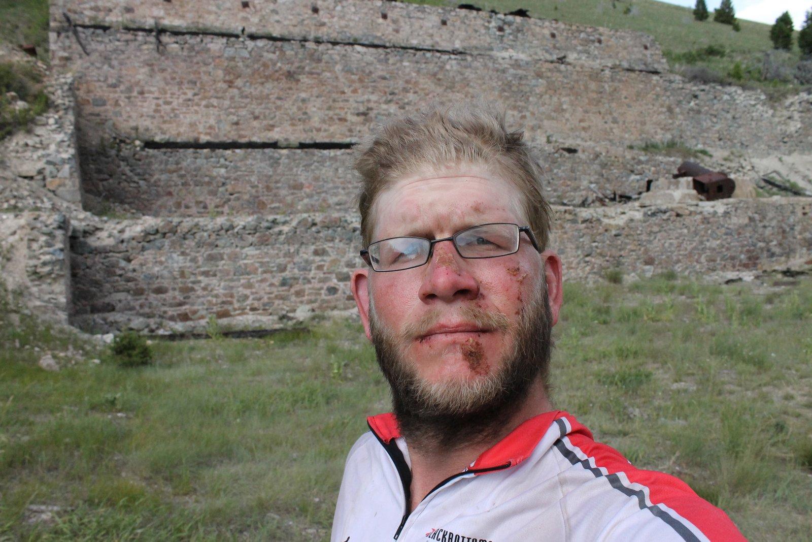 Dirt-and-blood-streaked rider in cycling gear posing in front of a stone mine ruin during the Great Divide route