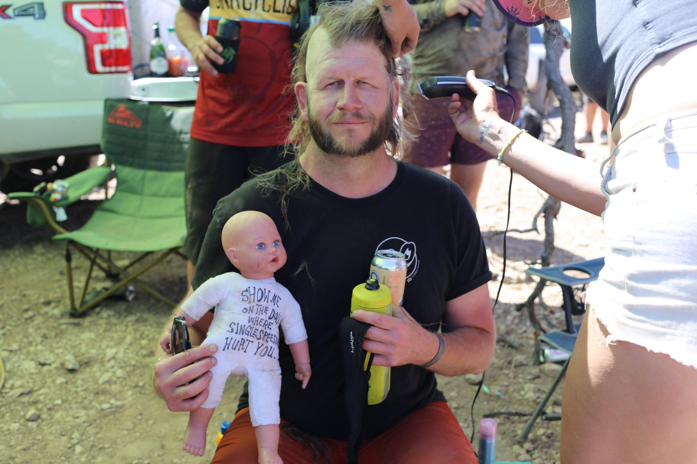 Bearded rider having his head shaved outdoors, holding a can and a baby doll with a shirt reading SHOW ME ON THE DOLL WHERE SINGLESPEED HURT YOU