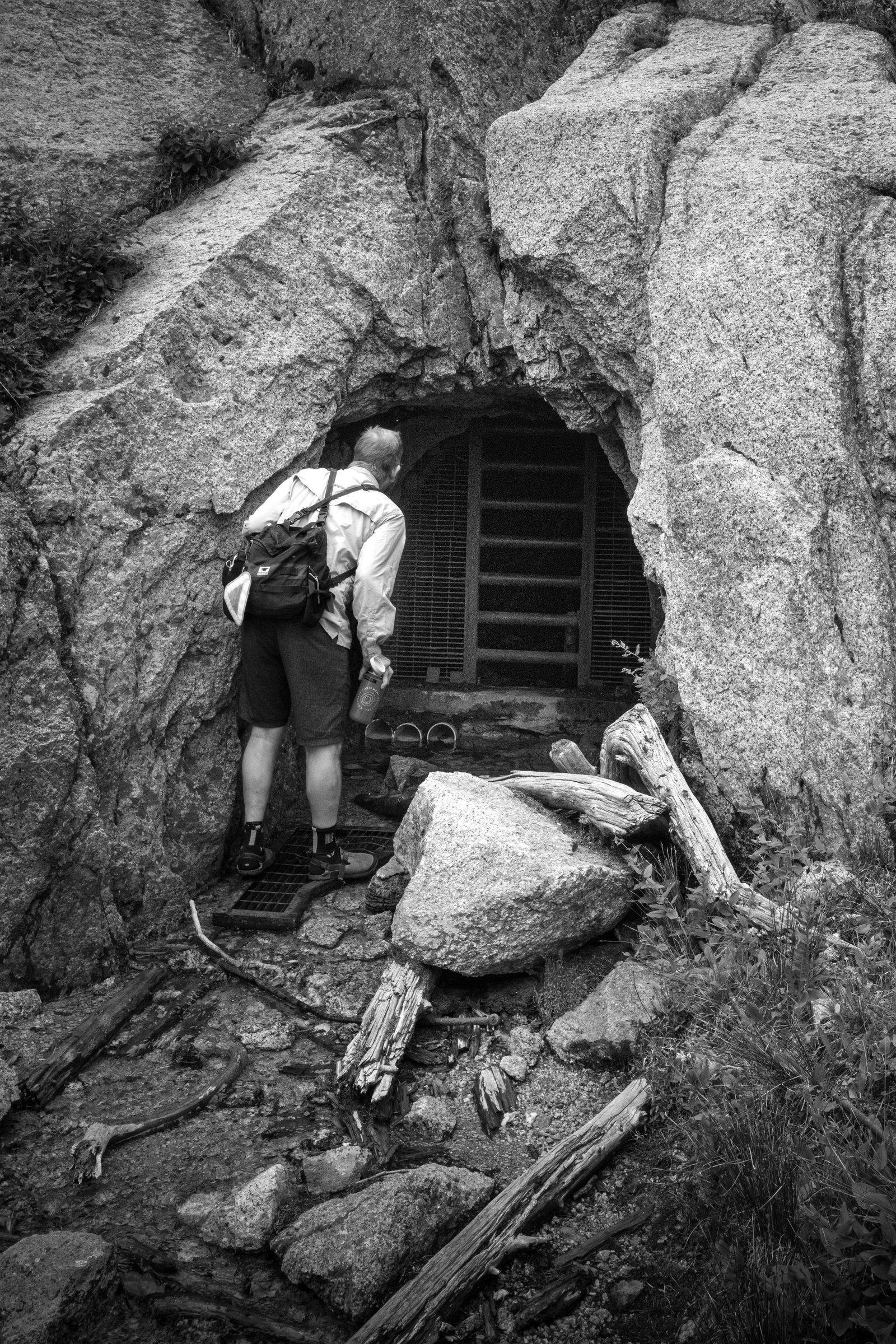 Black and white photo of a rider peering into a barred mine adit cut into a granite outcrop beside the trail
