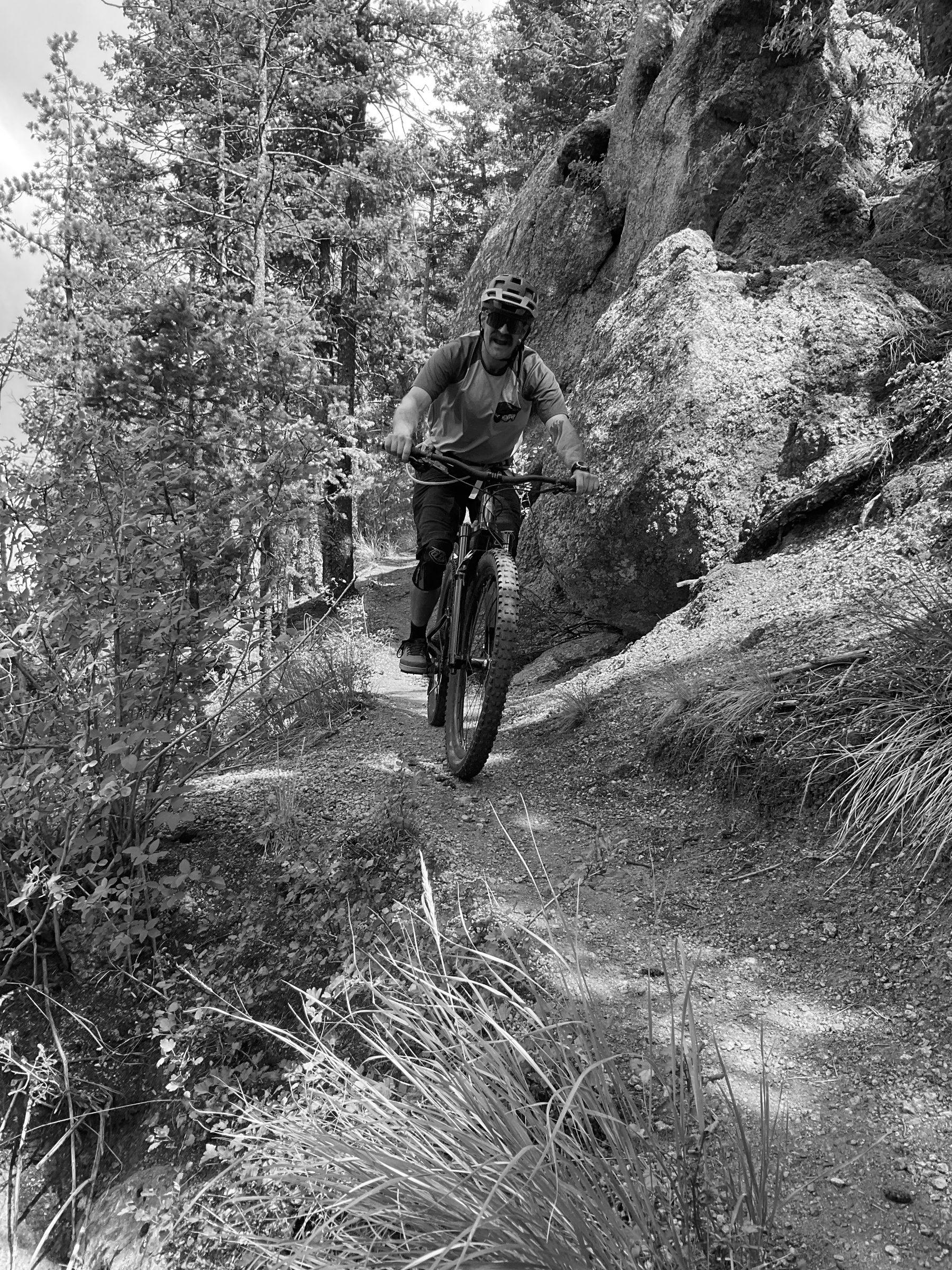Rider in a blue and gray jersey riding a fat-tire bike along narrow singletrack beside a large granite boulder