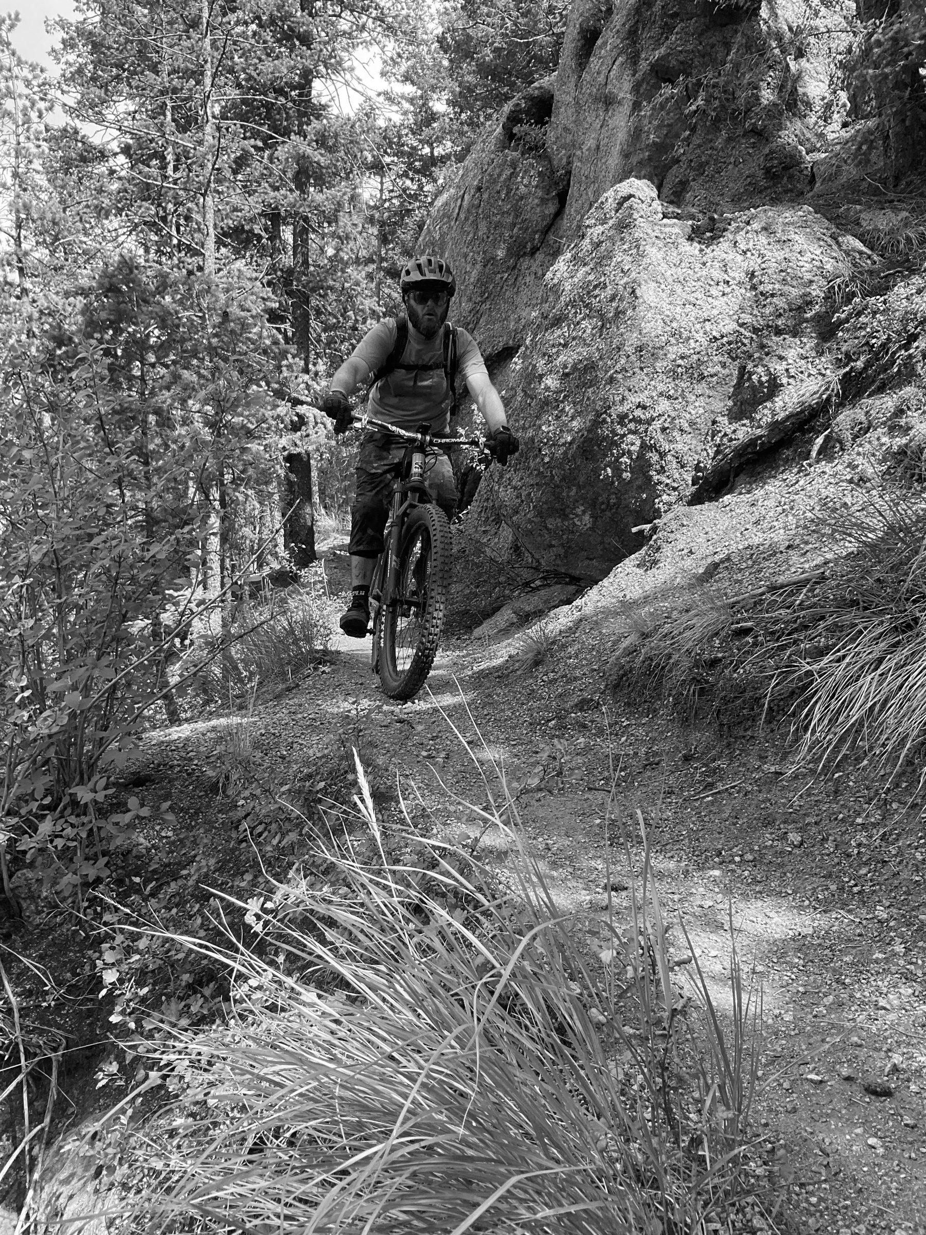 Rider in a mauve shirt riding a Maxxis-shod mountain bike on loose sandy trail below a granite outcrop