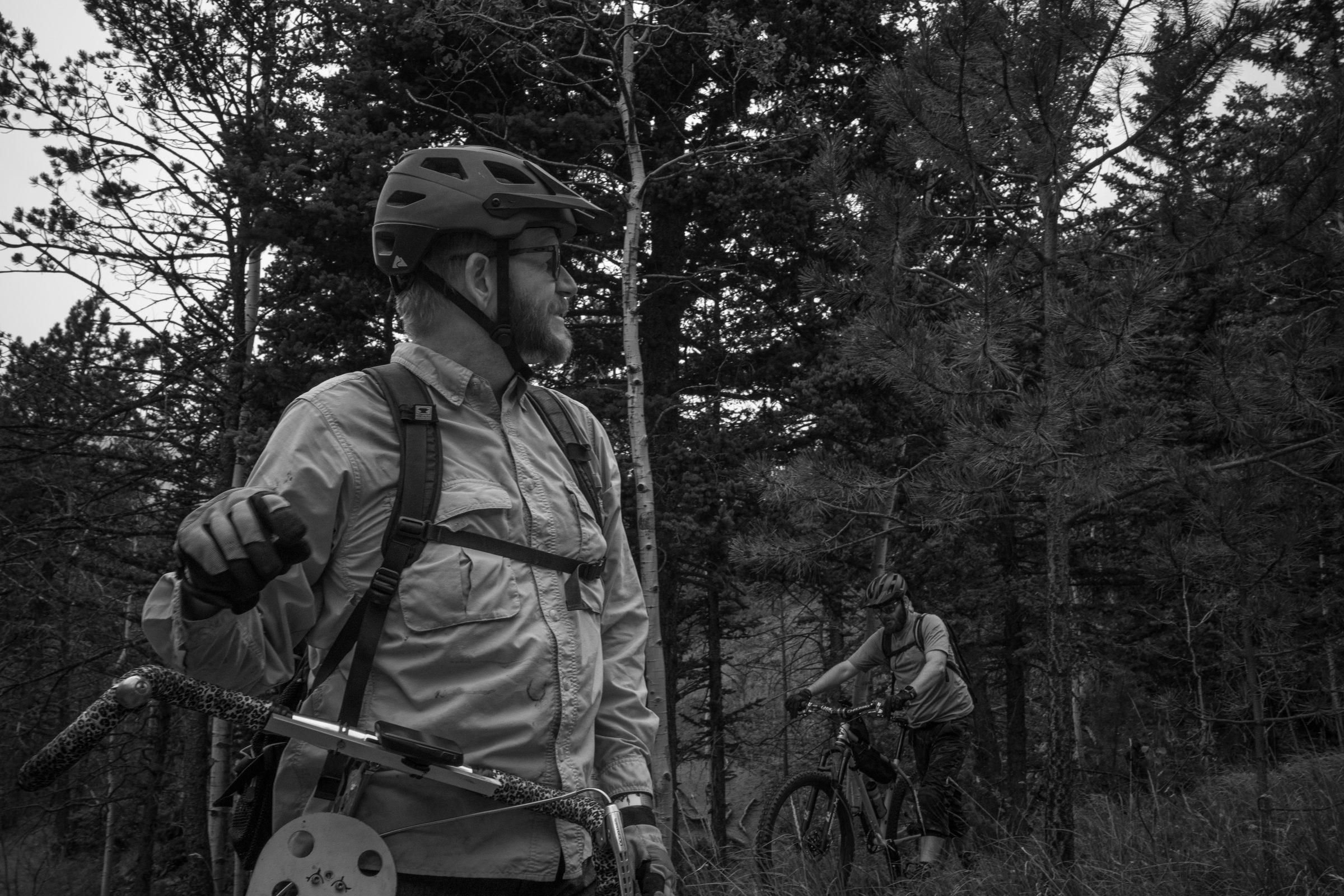 Black and white photo of Dave in the foreground with another rider behind him, both moving through a stand of aspens
