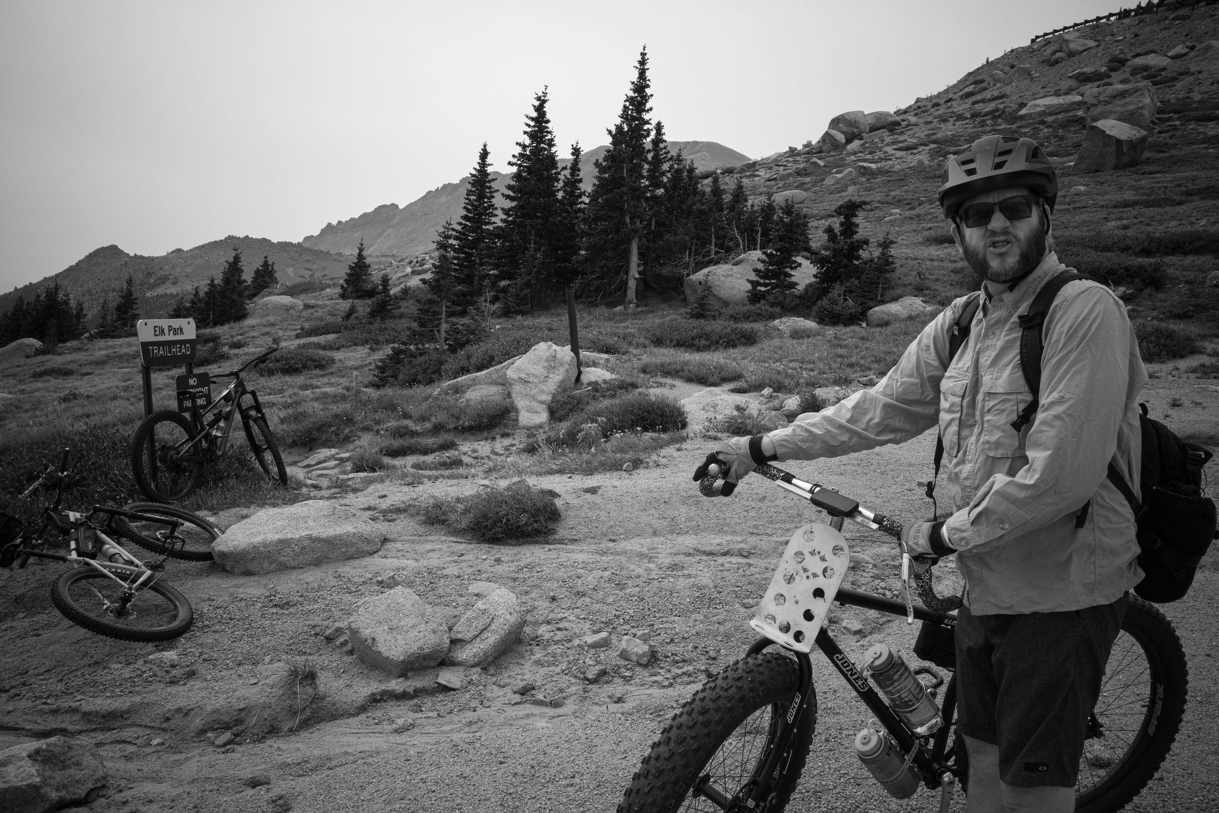 Black and white photo of the Elk Park trailhead sign with three bikes leaning nearby and peaks rising behind