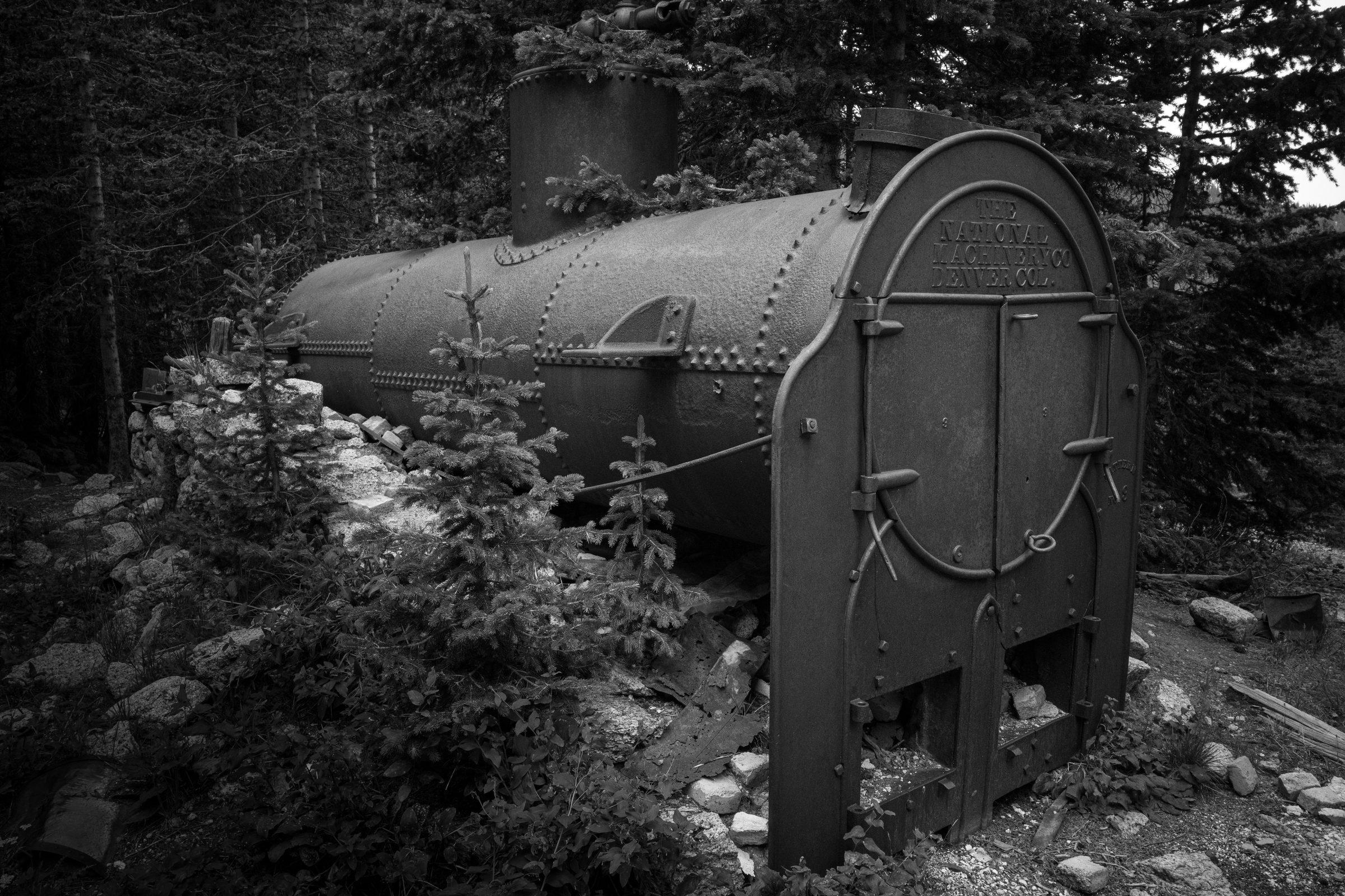Black and white photo of a rusted steam boiler stamped National Machinery Co Denver, backed by spruce
