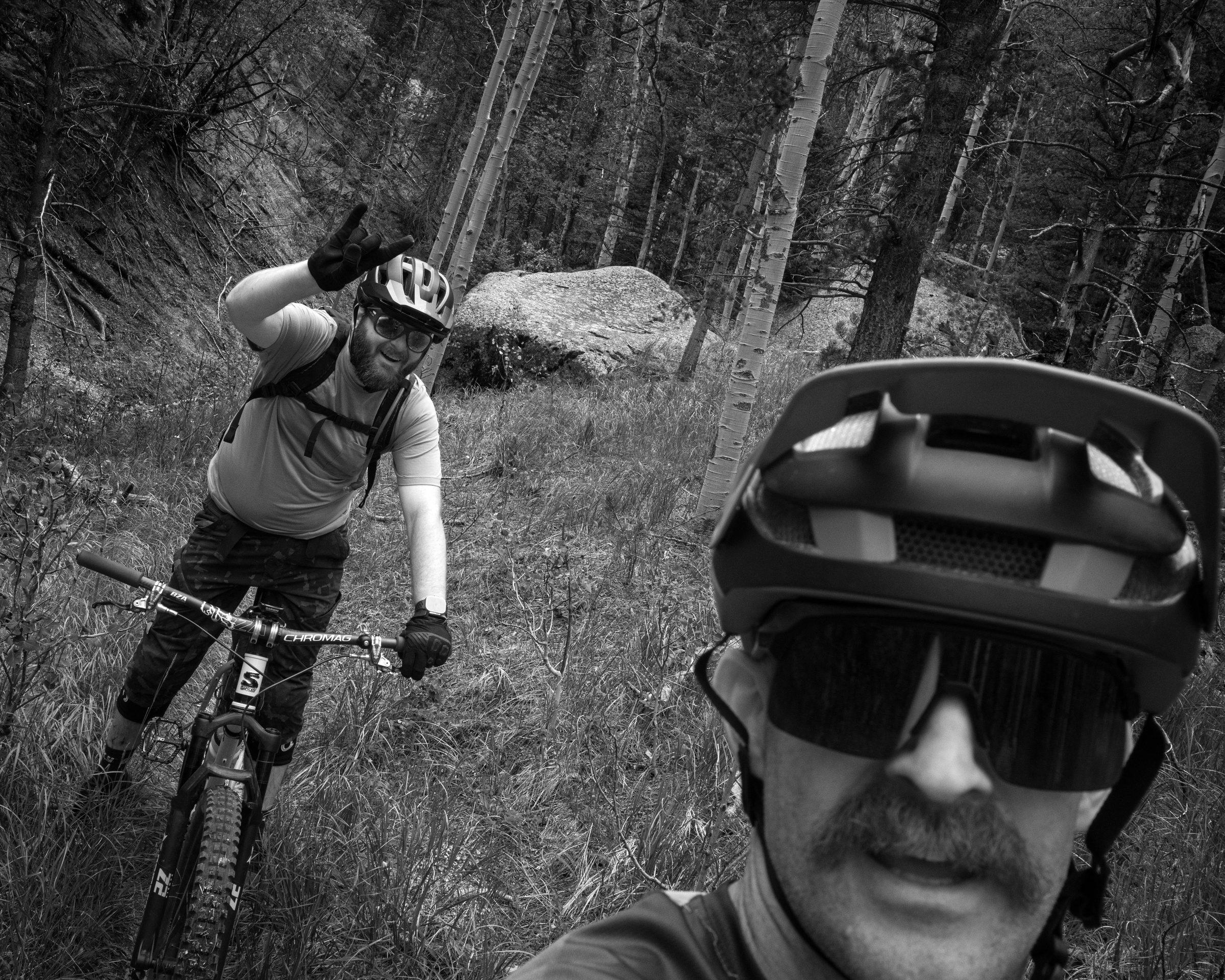 Black and white selfie of a rider up front with another rider throwing a peace sign behind, mountains beyond