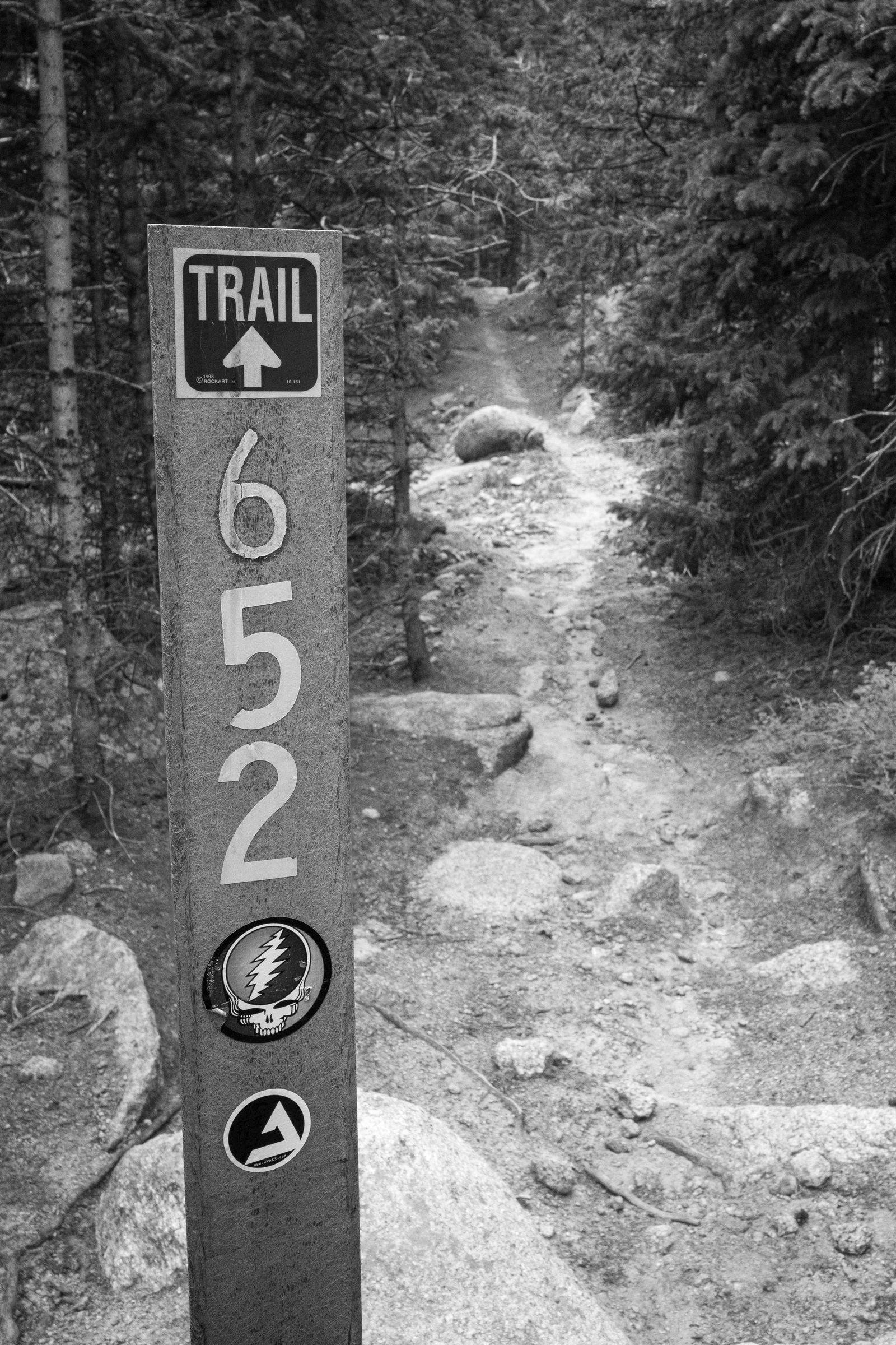 Black and white photo of a trail marker post reading TRAIL 652 with a Grateful Dead sticker, rocky singletrack dropping into the trees behind