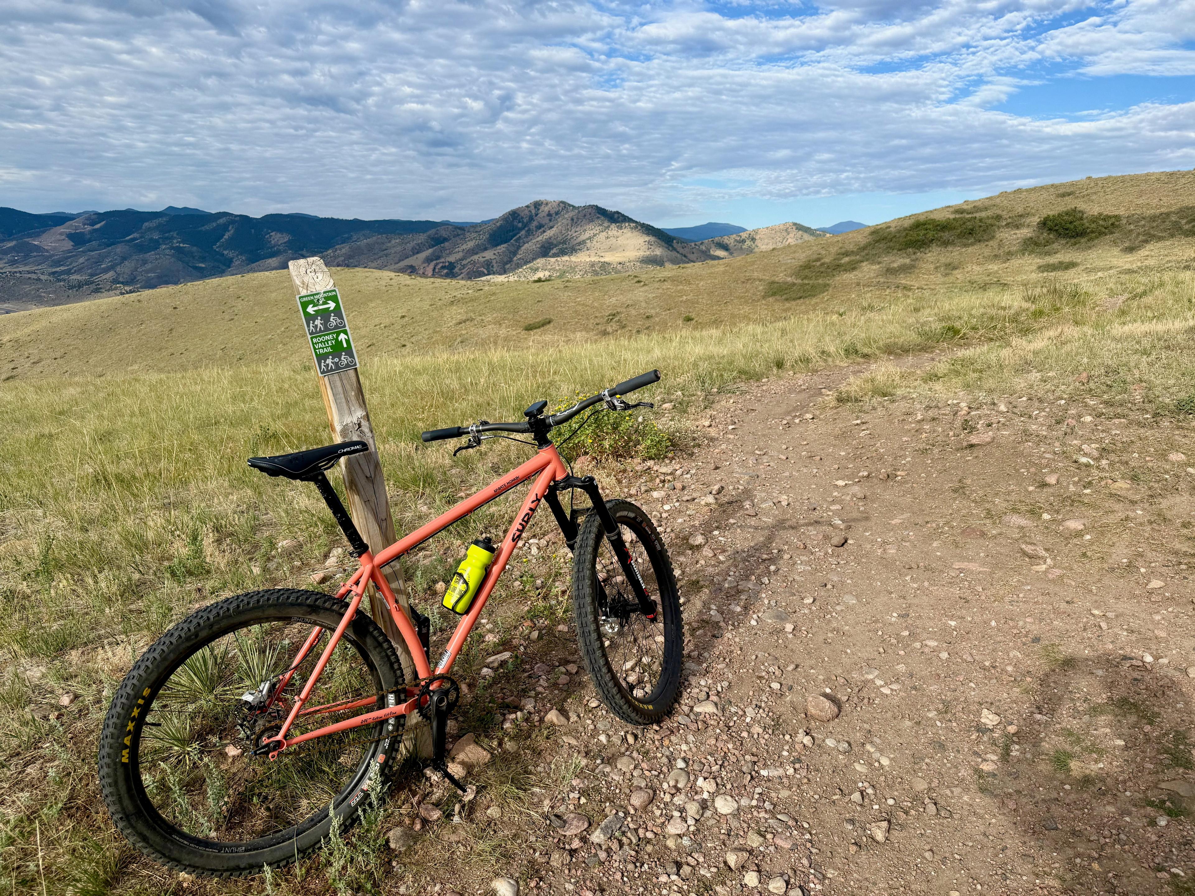 Salmon Surly Karate Monkey leaning against a Green Mountain Trail and Rooney Valley Trail sign on open, exposed grassland with foothills behind