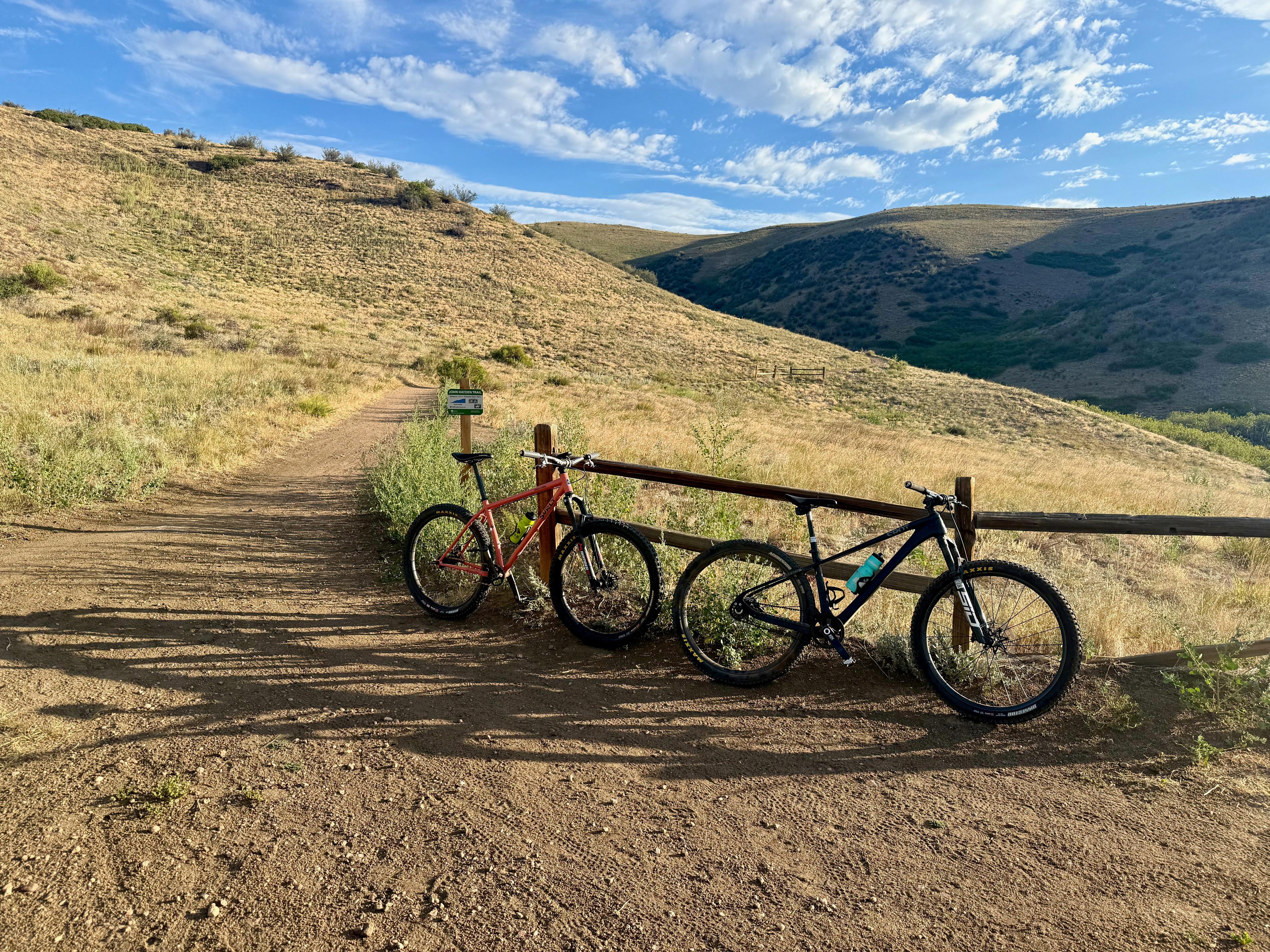 Two mountain bikes leaning on a split-rail fence beside a green John Hayden Trail sign, with dry grassland hills and a shaded valley behind