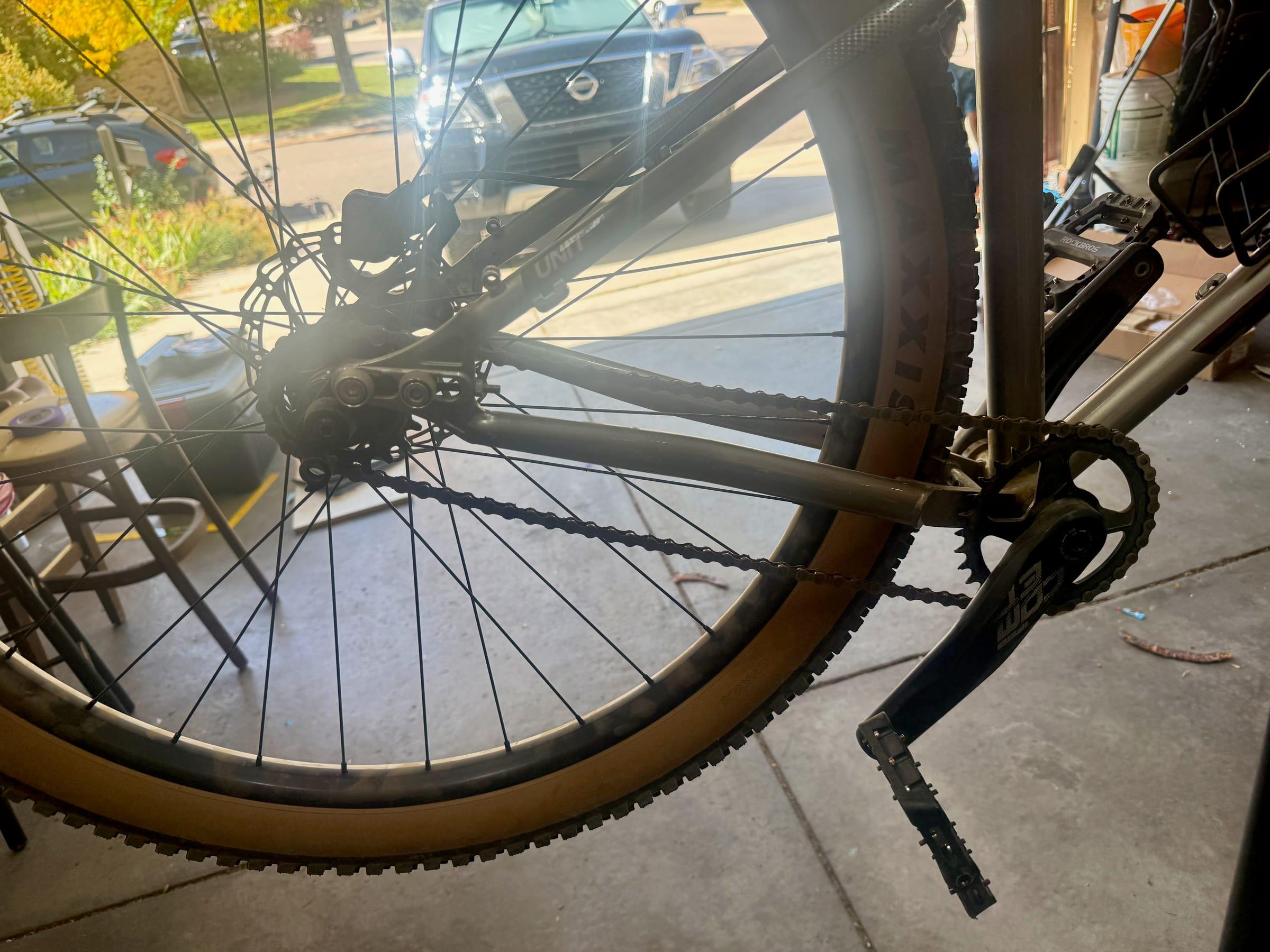 The drive side of a Kona Unit on a repair stand, showing a black FSA crank arm with a flat pedal, a single chainring, and the chain running back along the chainstay to the rear wheel
