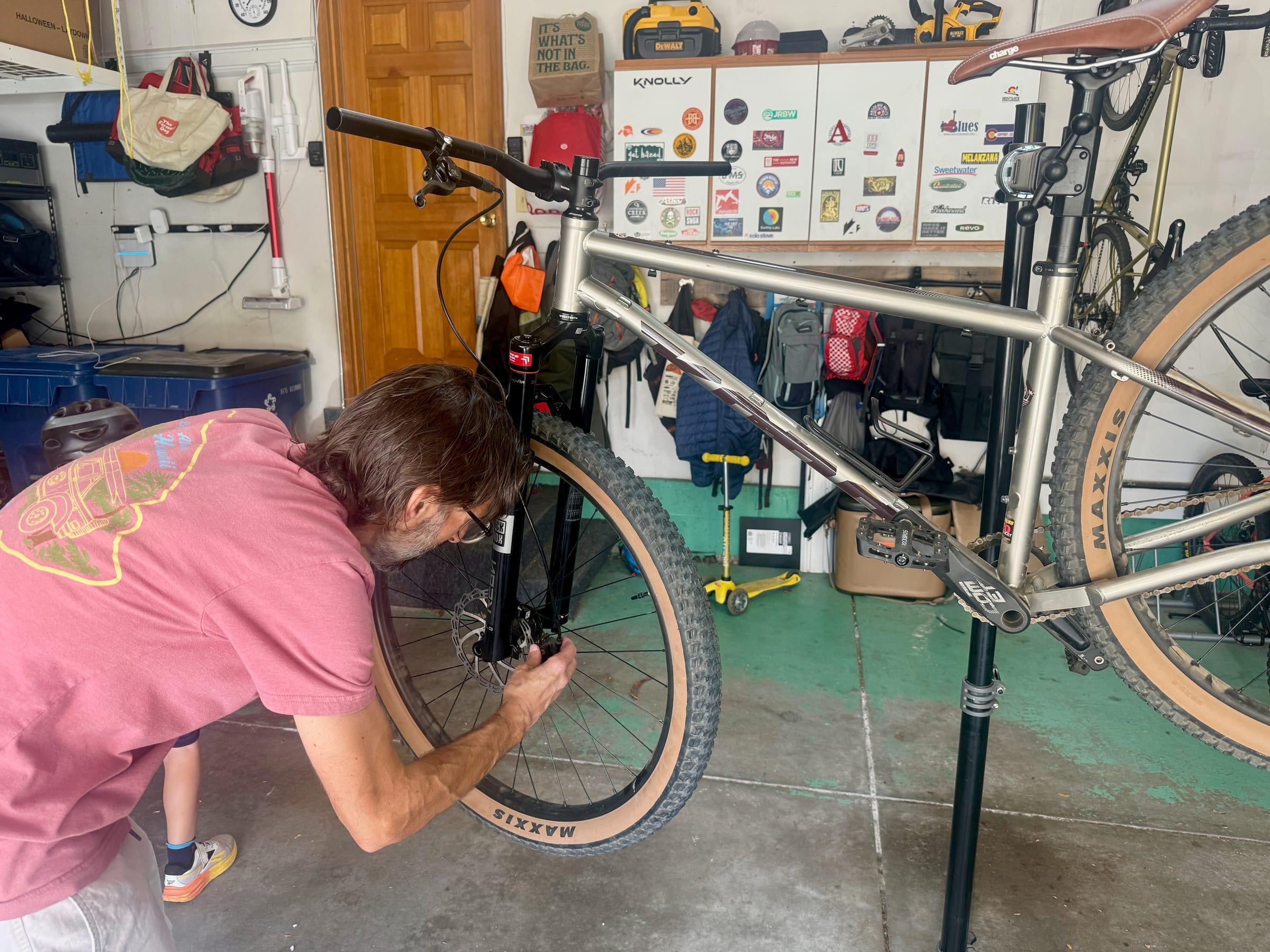 Brad kneeling beside the front wheel of a Kona Unit with a black RockShox Reba fork, working on the front brake caliper, with a brown Charge saddle on the bike next to him in the garage