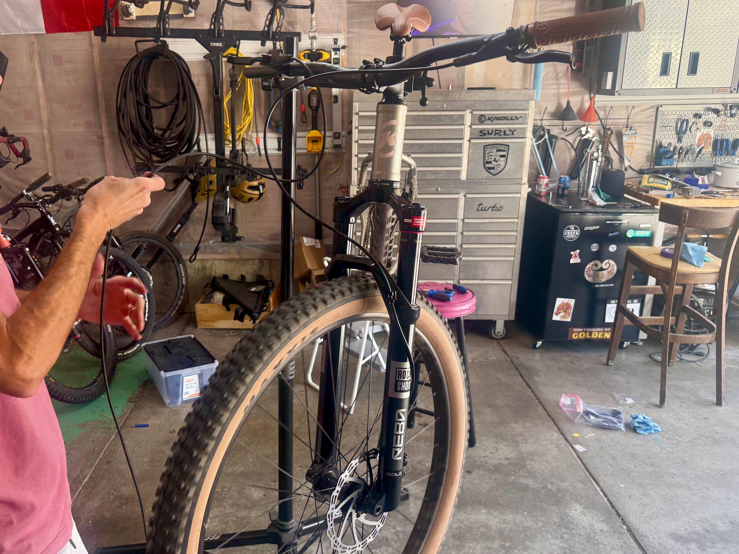 A Kona Unit on a repair stand in a garage, seen from the front, with a RockShox Reba fork and a tan-wall Maxxis tire, a long brake hose looping across the handlebars, and a hand holding the end of the hose
