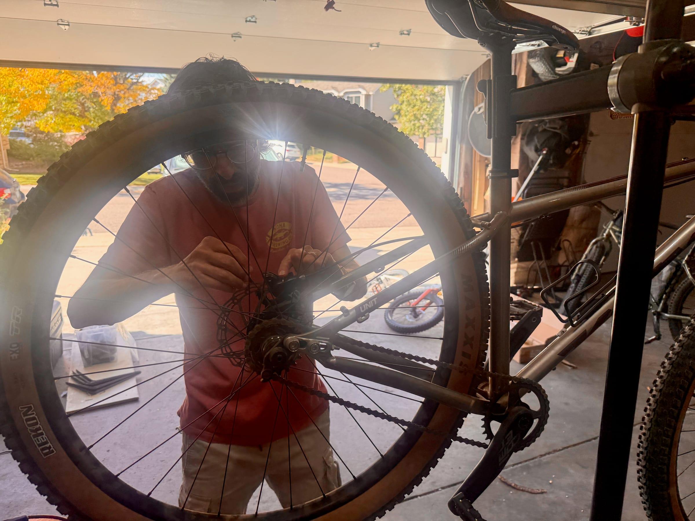Brad working on the rear brake caliper of a Kona Unit, seen through the spokes of the rear wheel in a garage with a cog and chain visible below