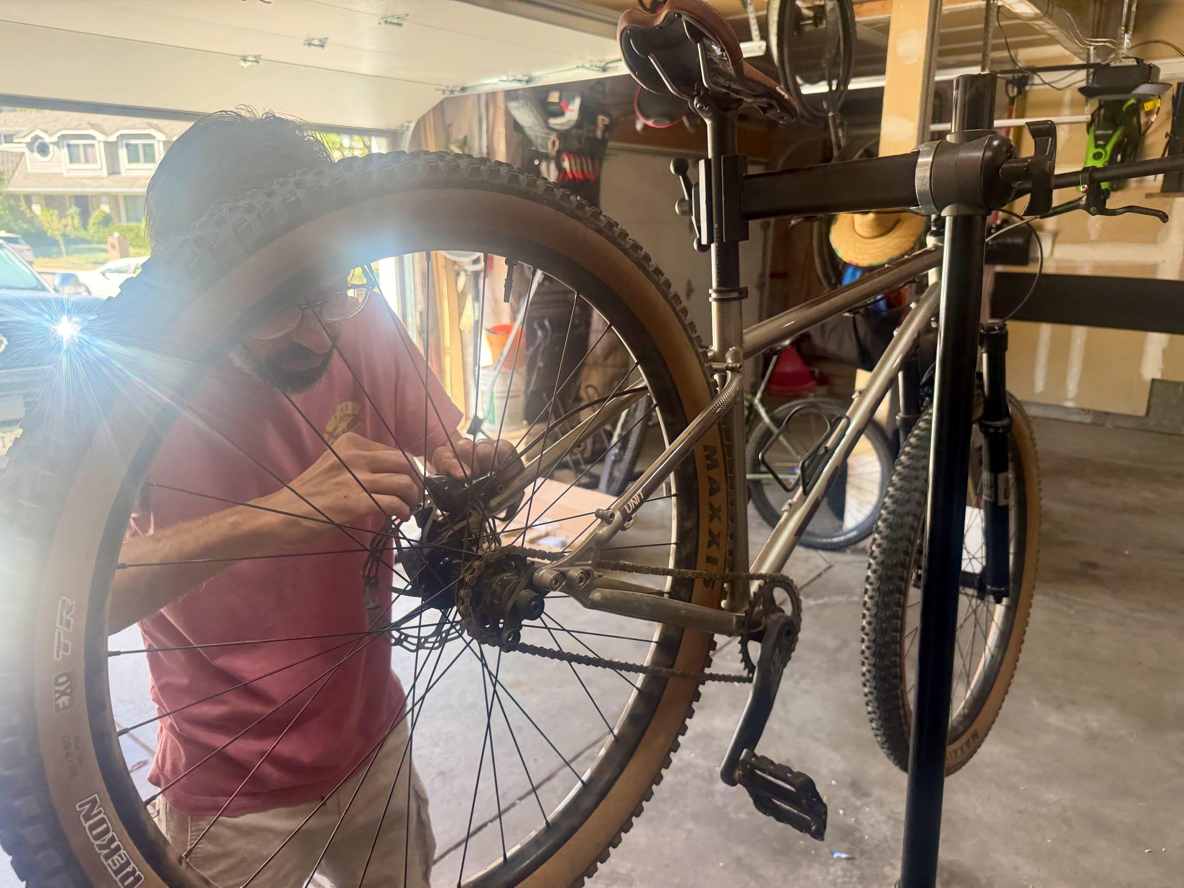 Brad leaning in to work on the rear hub of a Kona Unit, seen through the spokes of the rear wheel, with the cog and chain visible next to the rear brake caliper