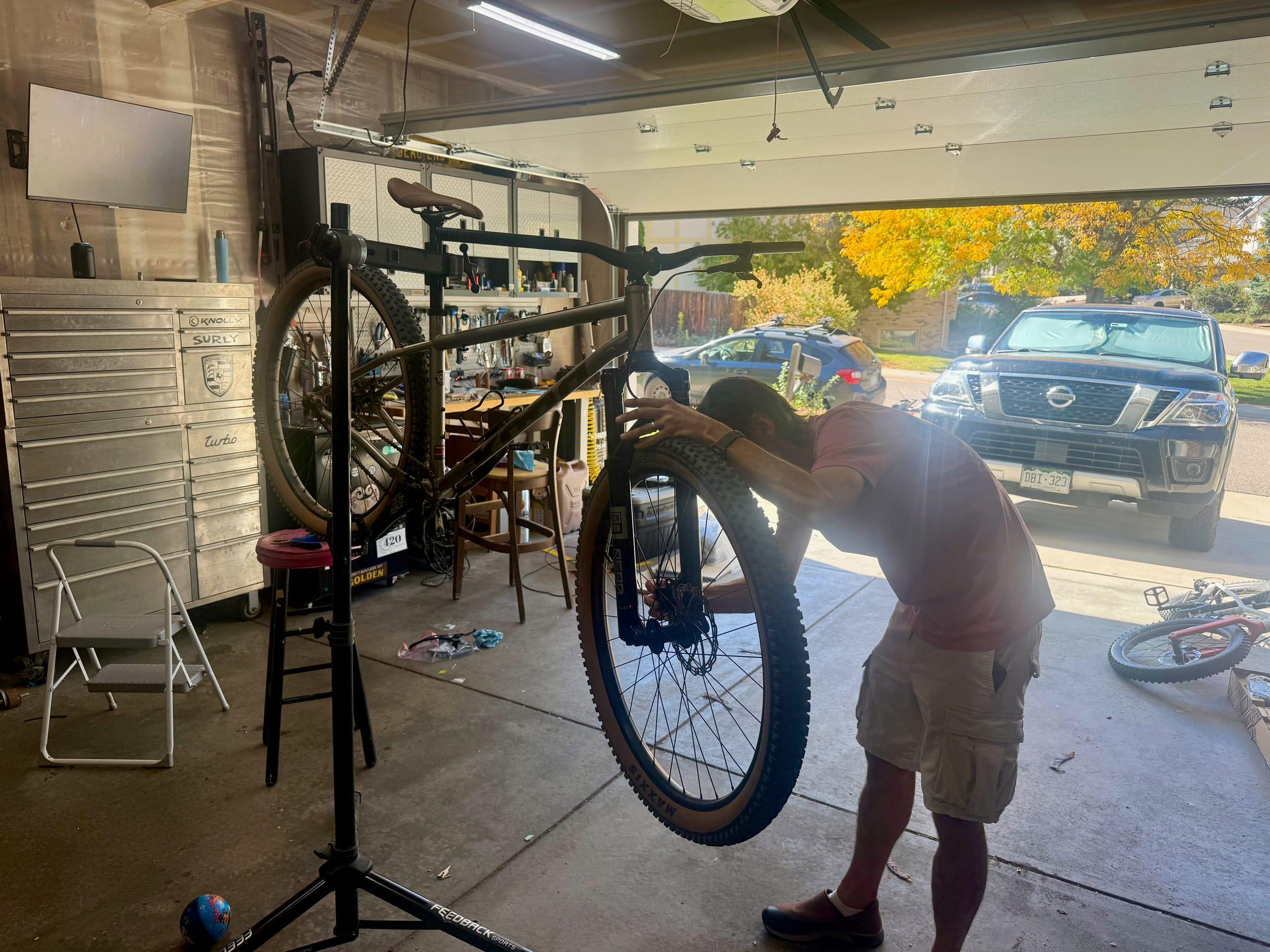 Brad bent over a front wheel on a Kona Unit mounted in a repair stand in a garage, with tan-wall Maxxis tires, an open garage door, and a black Nissan Armada parked in the driveway