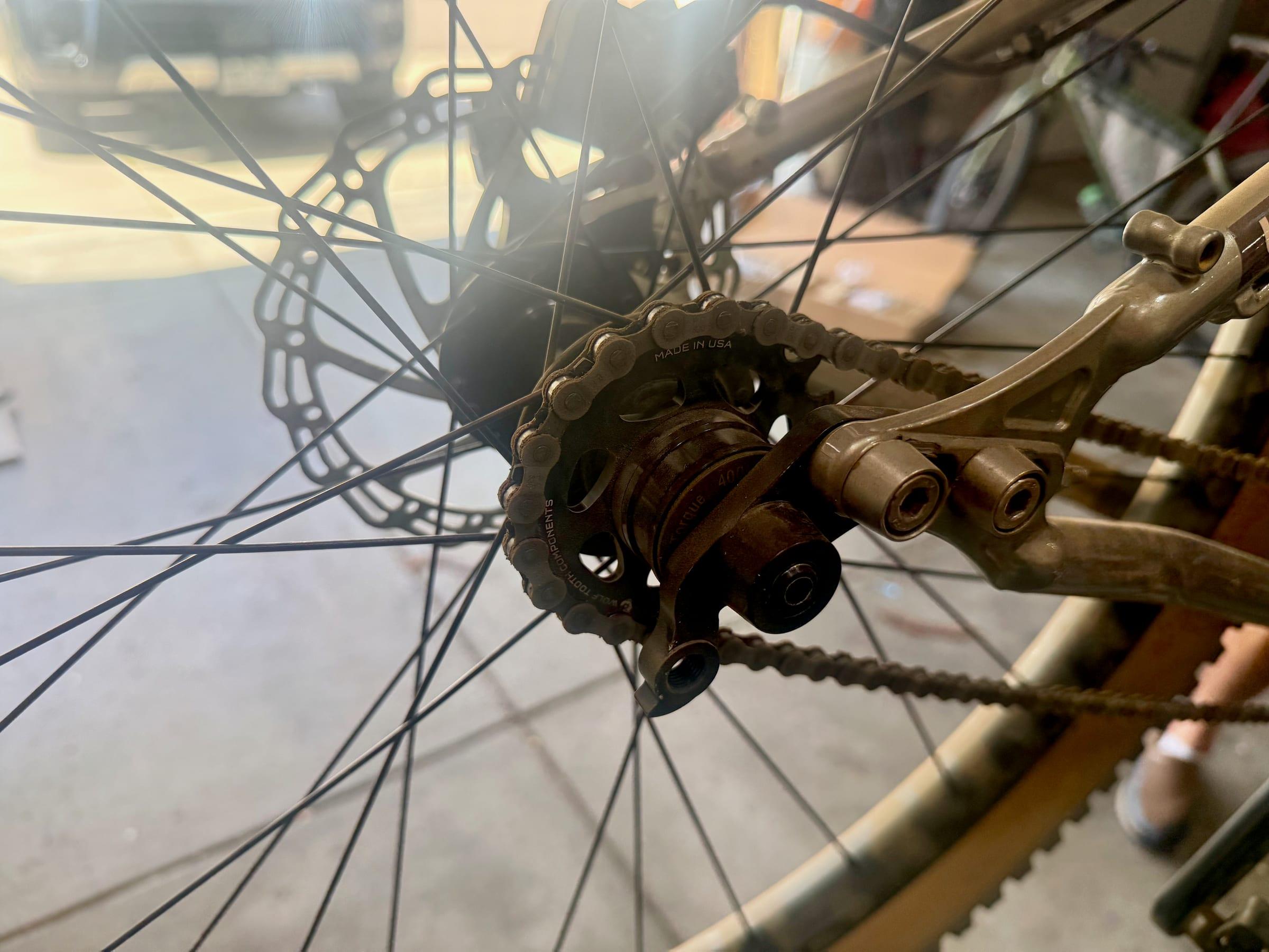 Close-up of a black Wolf Tooth cog on a rear hub with a stack of spacers beside it, the chain wrapped around the cog, and the brake rotor and dropout behind it