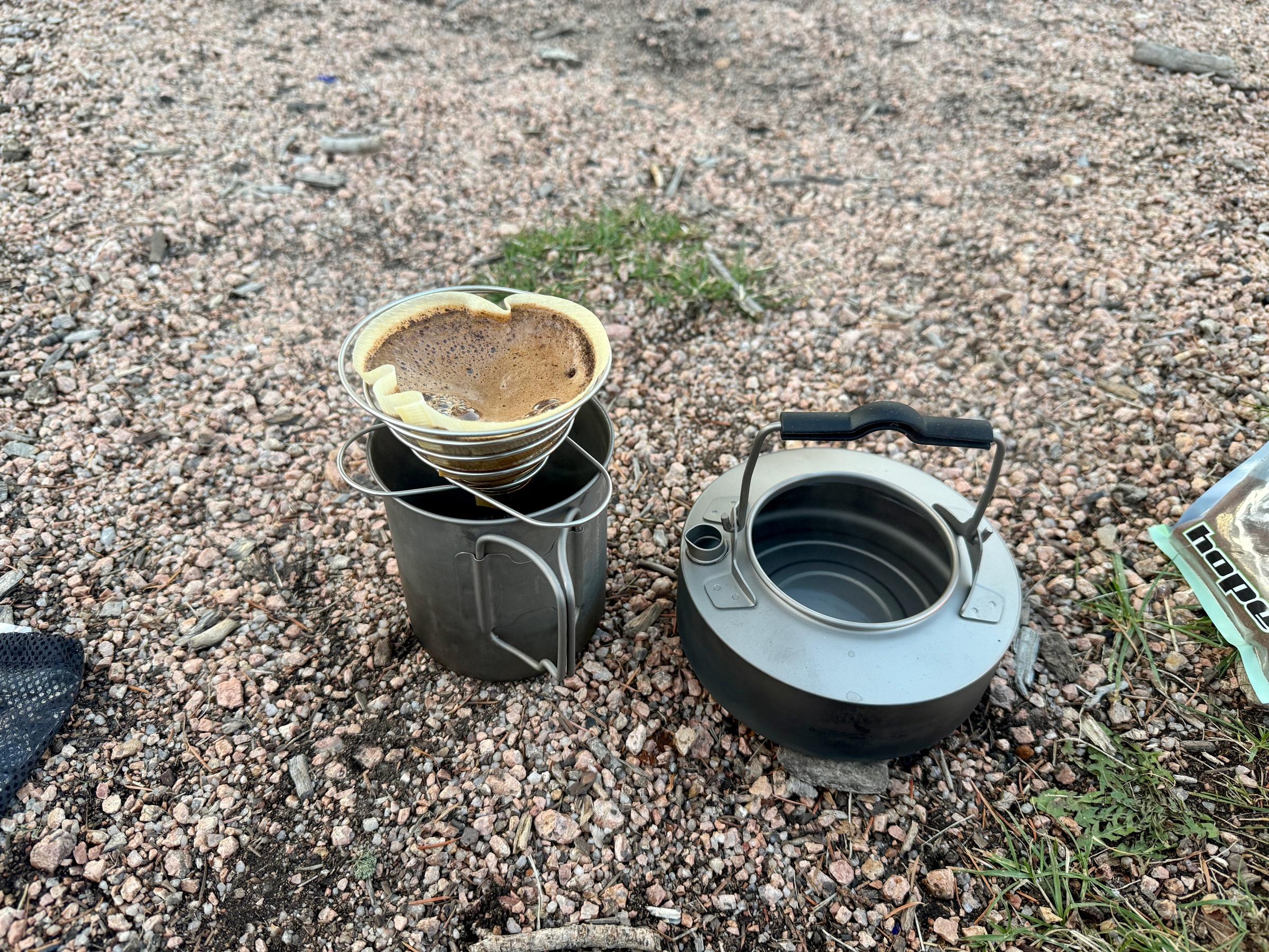 SOTO Compact Helix coffee dripper with a used paper filter sitting atop a titanium cup, kettle beside it on gravel