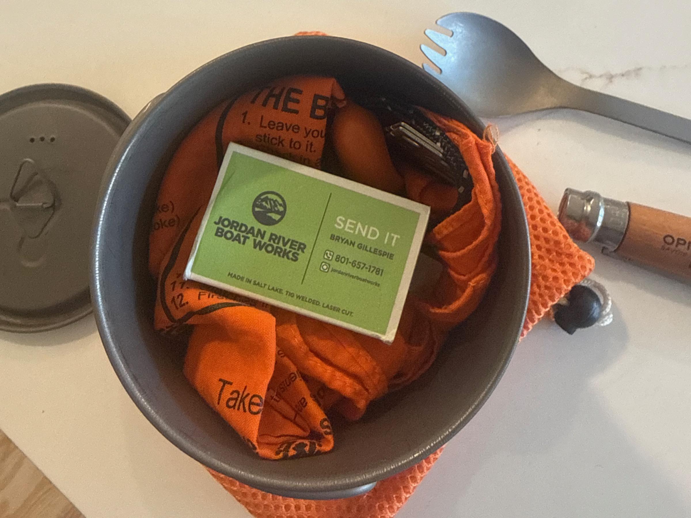 The old Backcountry titanium cup with its lid off, packed with a folded orange bandana and a small knife, the Trangia burner's cap and a titanium spork resting beside it
