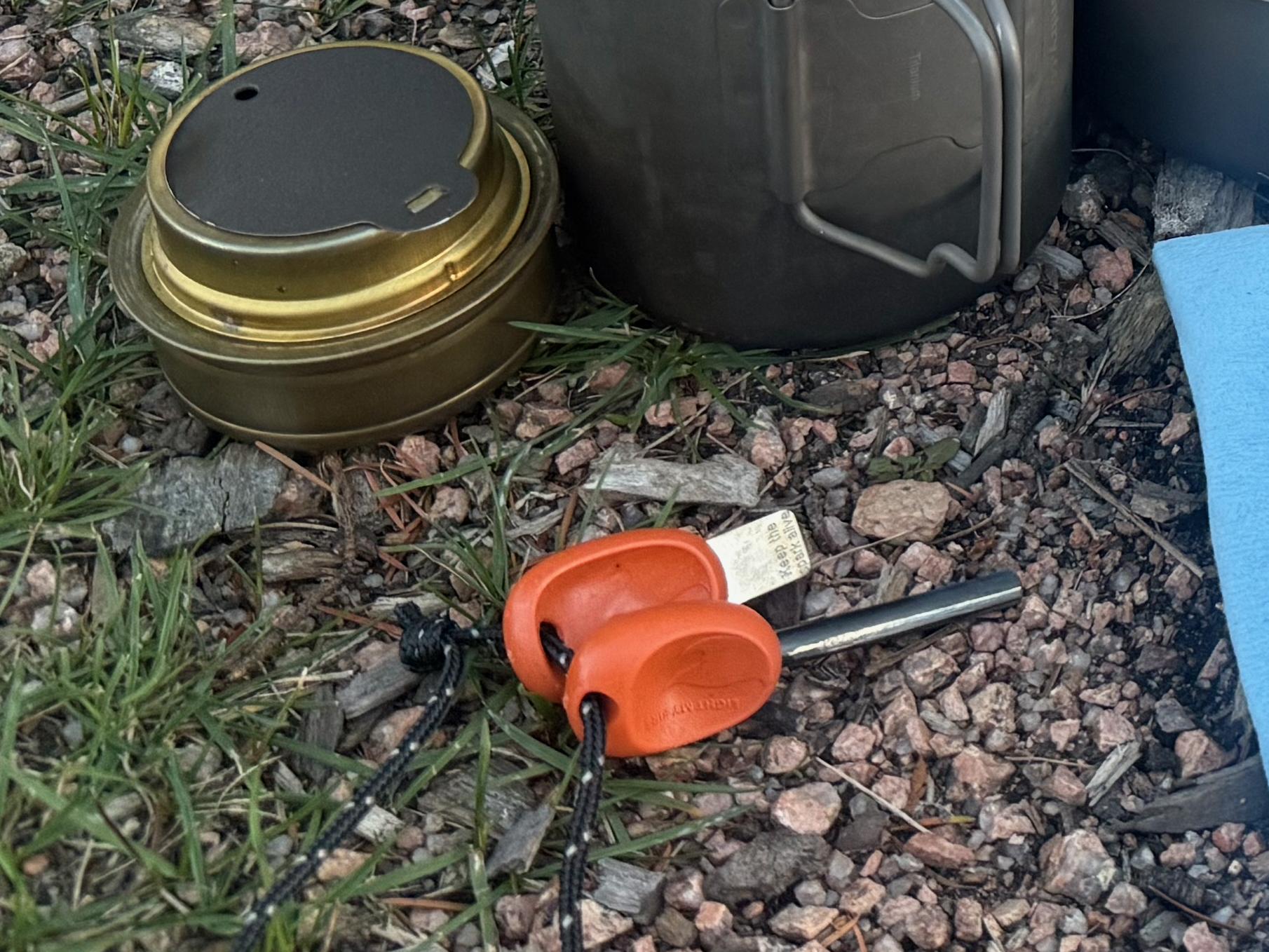 Light My Fire FireSteel ferro rod resting next to the Trangia burner's brass cap on gravel and grass