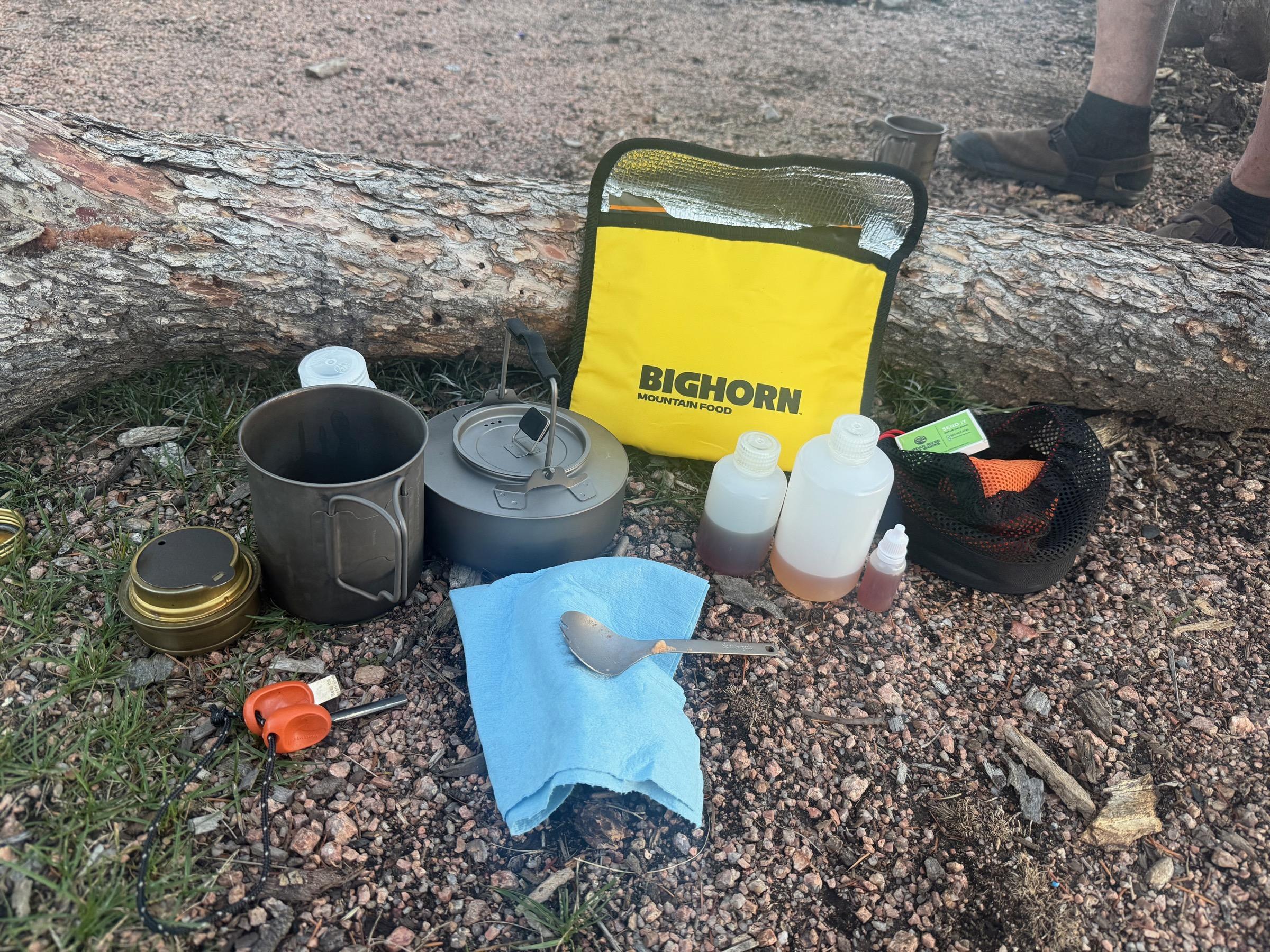 The complete bikepacking cooking kit laid out against a fallen log: BIGHORN insulated bag, titanium kettle and cup, Trangia burner, fuel bottles, mesh accessory bag, and a spoon resting on a blue bandana