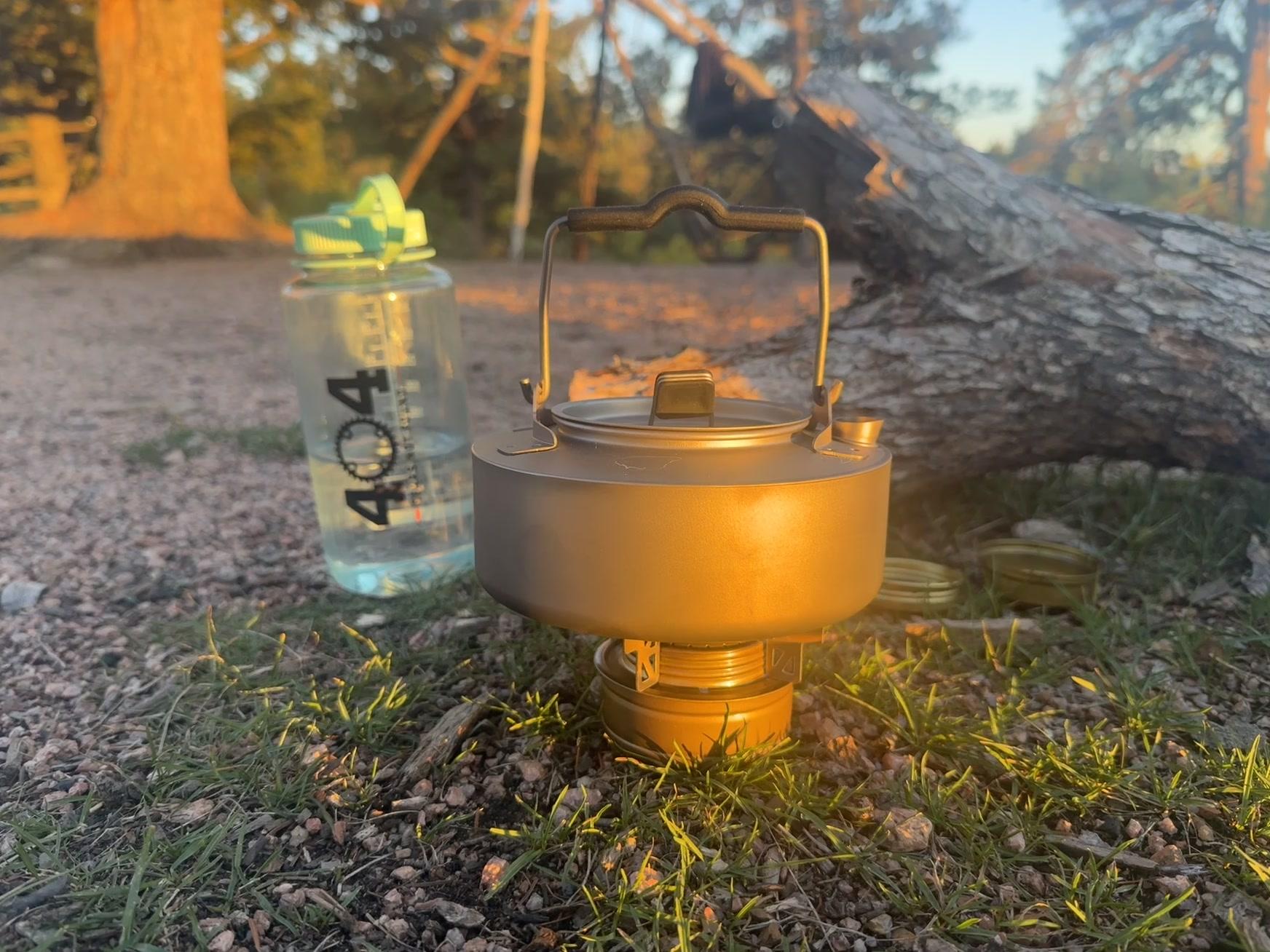 The titanium kettle glowing on the lit Trangia burner at golden hour, flame visible beside it, a 404 Gears Not Found water bottle in the foreground