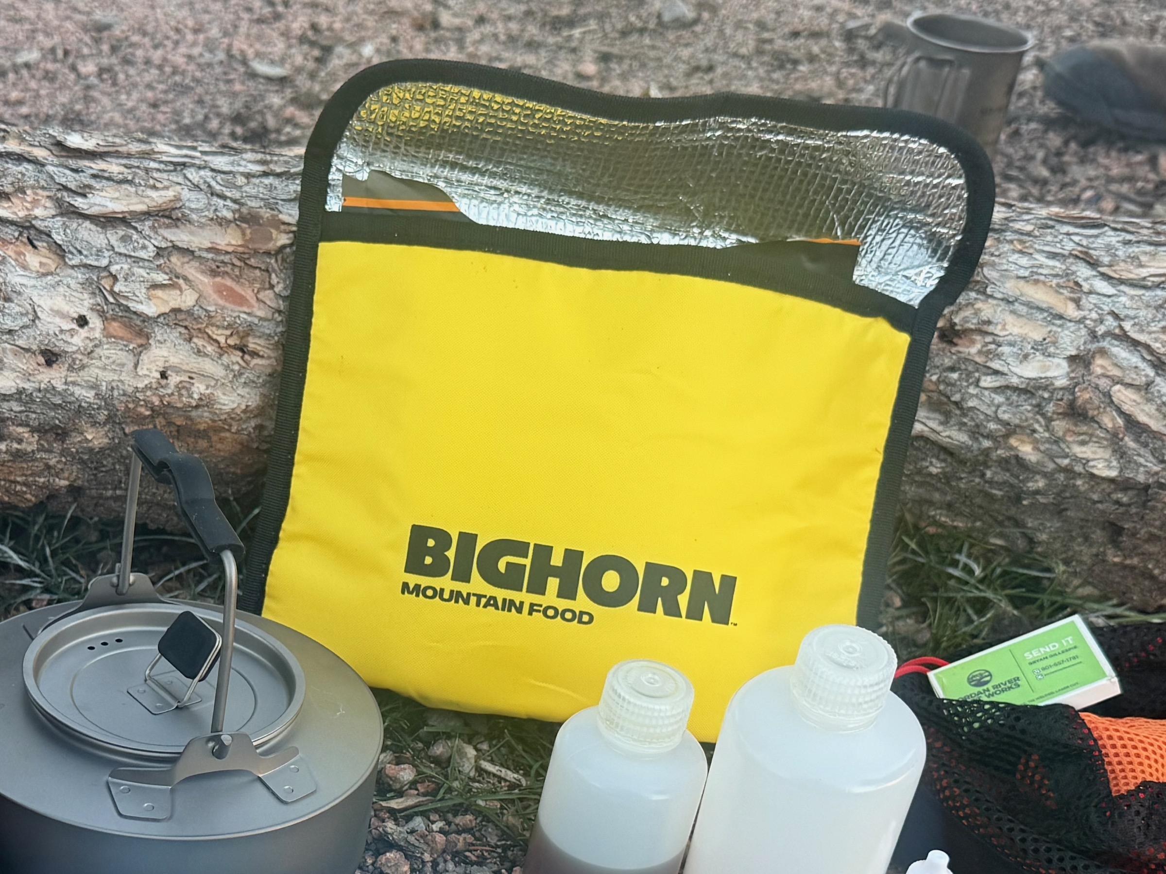 BIGHORN insulated bag standing open against a fallen log, foil-lined interior visible, kettle and fuel bottles beside it at camp