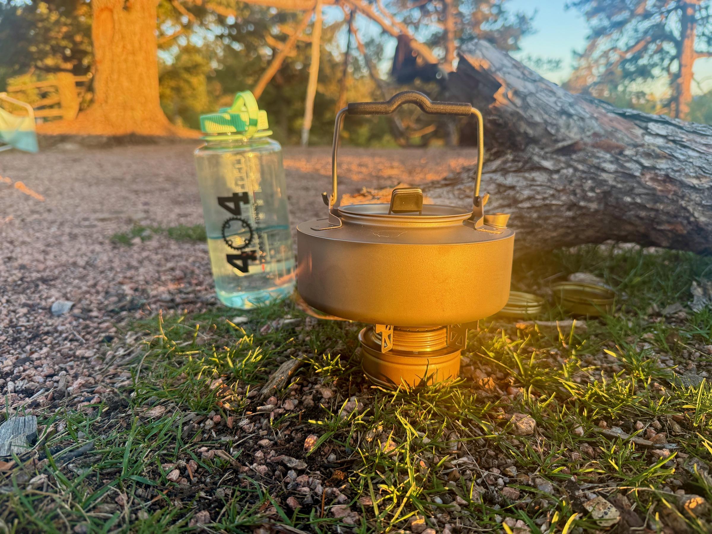 Boundless Voyage titanium kettle with its folding handle raised, sitting on the lit Trangia burner at golden hour