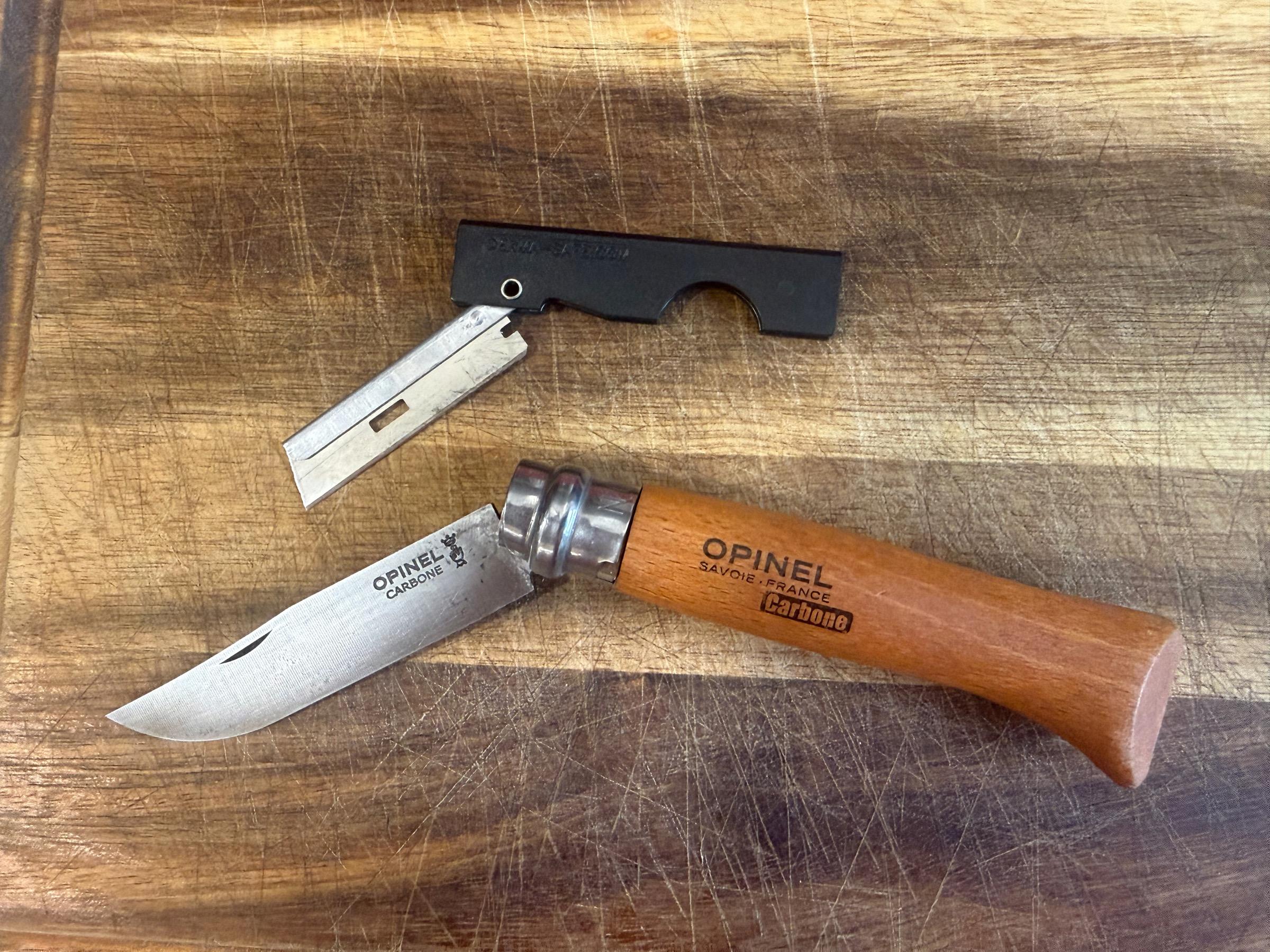 Derma-Safe folding utility knife open beside an Opinel No.08 Carbone with its beechwood handle, both resting on a well-worn cutting board