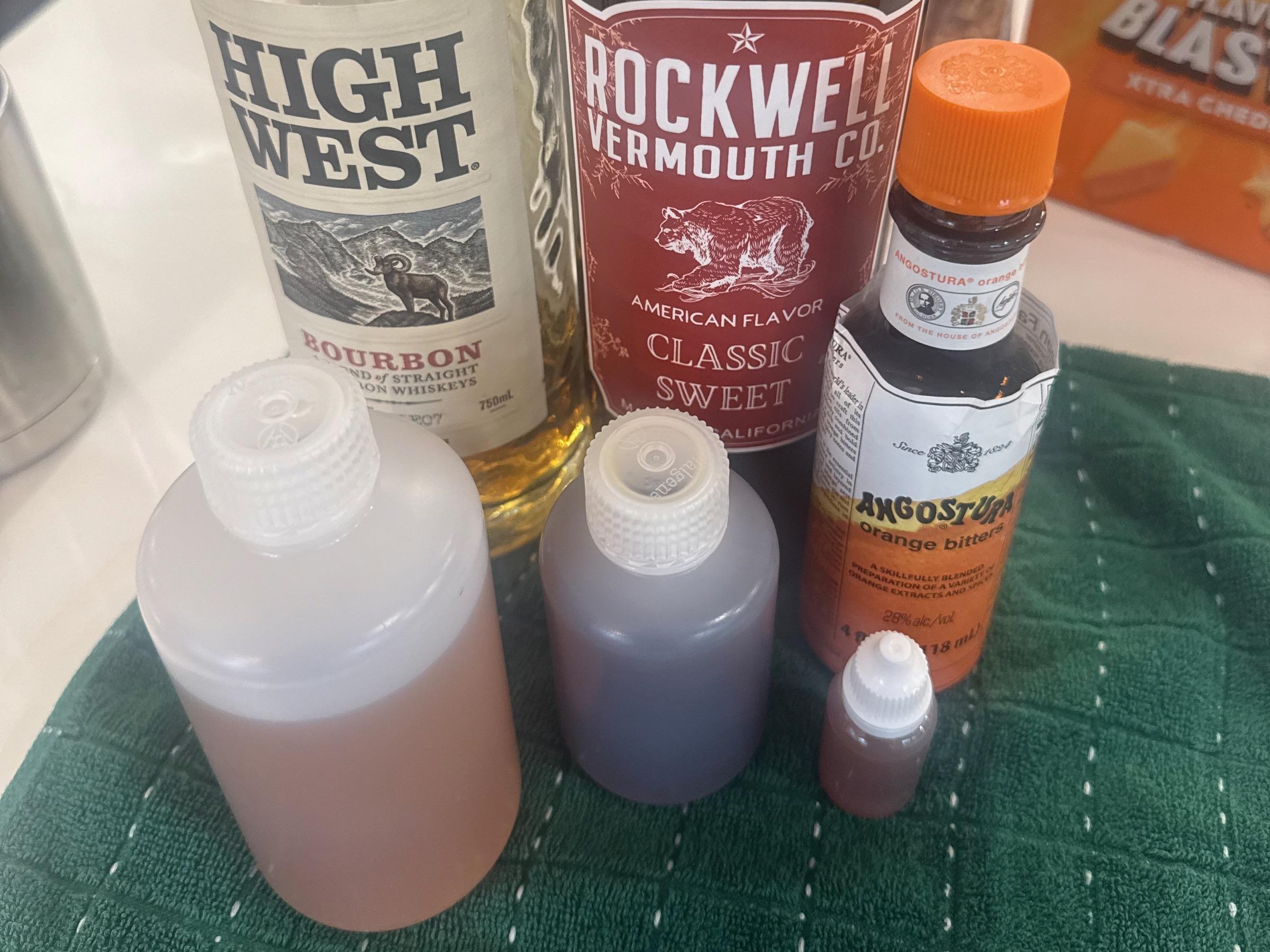 Small narrow-mouth Nalgene bottles filled with bourbon and vermouth, plus a small dropper bottle of orange bitters, posed next to the actual liquor bottles they came from