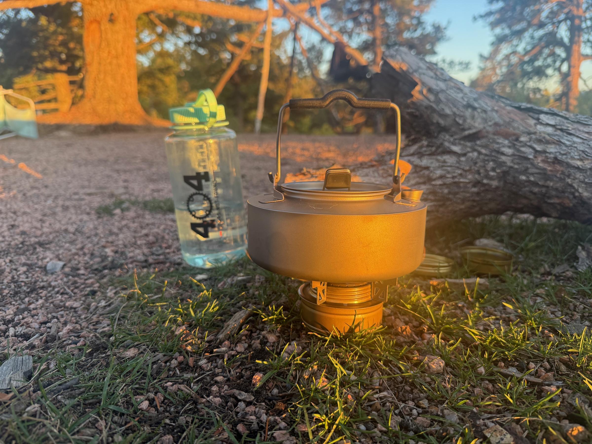Trangia alcohol spirit burner assembled beneath the titanium kettle at golden hour, a 404 Gears Not Found water bottle beside it