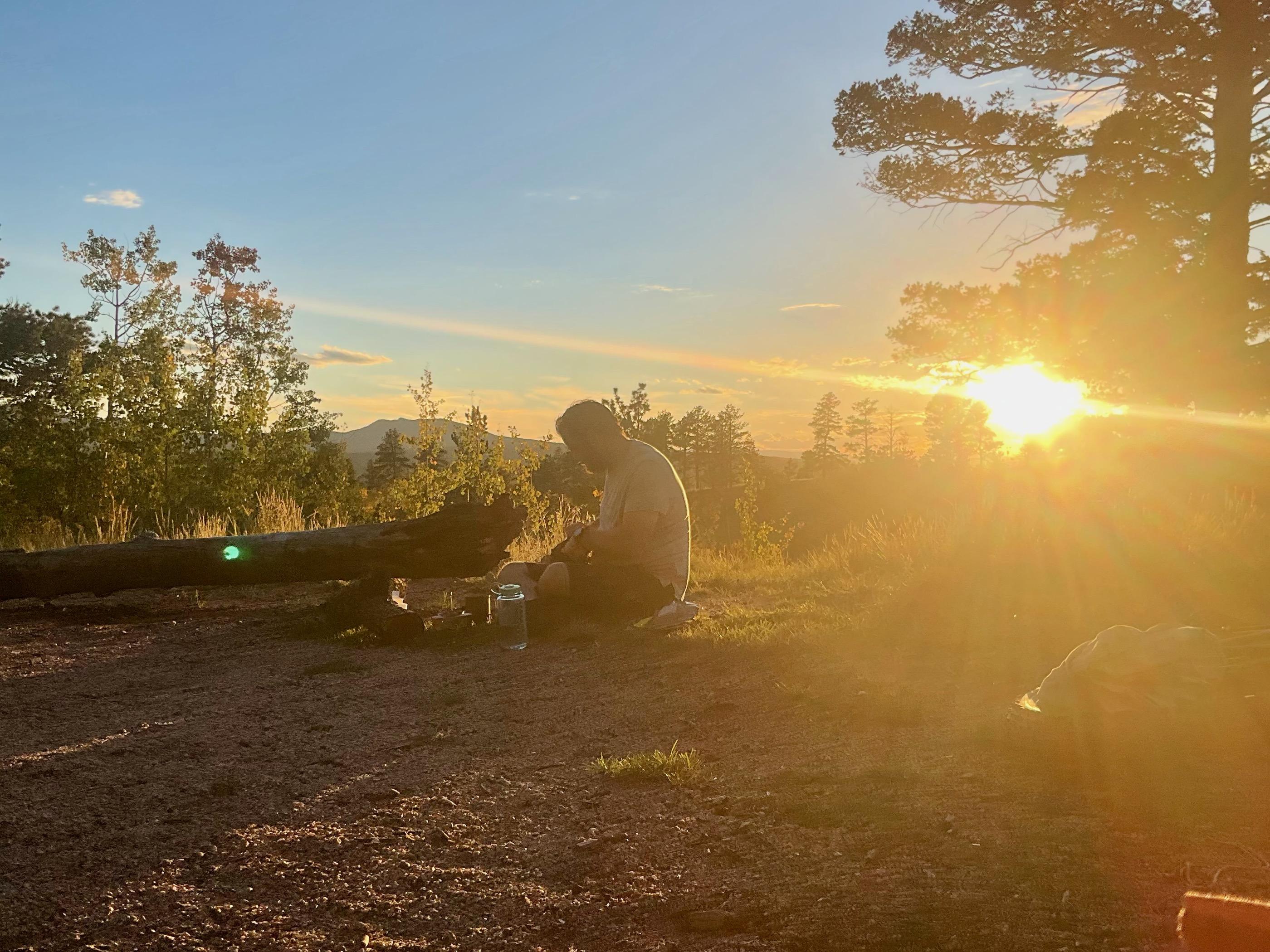 A silhouetted figure crouched over a camp stove at sunset beside a fallen log, sun flaring through the trees behind him
