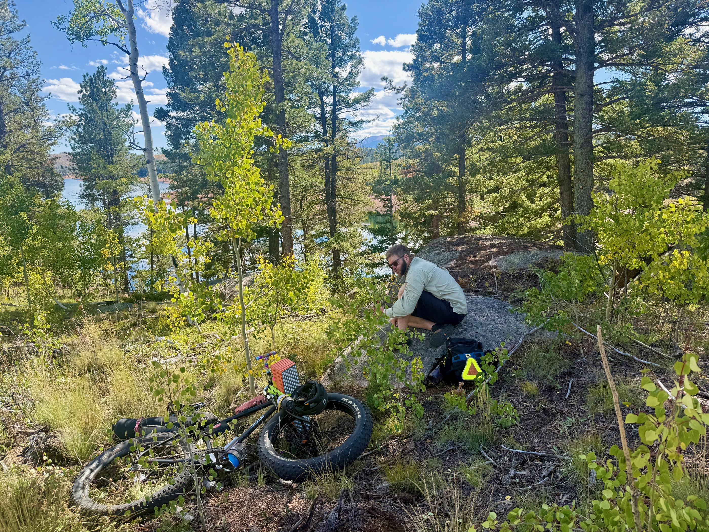 Fixie Dave crouched beside his loaded fat bike on a rocky outcrop above the reservoir, forest and water behind him