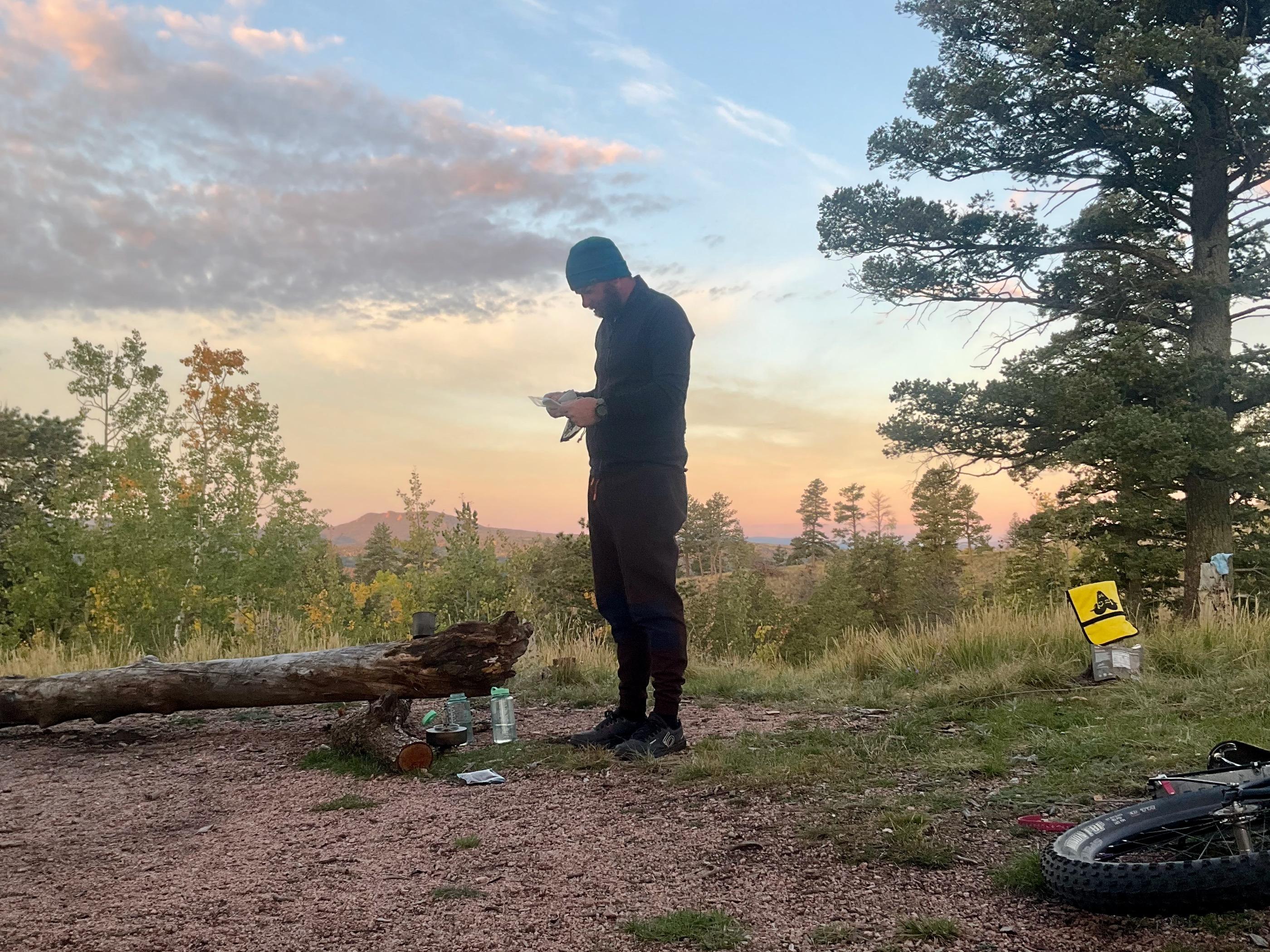A rider standing at the dispersed camp at dawn sorting gear near a fallen log, a small 404 Gears Not Found flag hanging from a branch, fat bike tire visible in the foreground