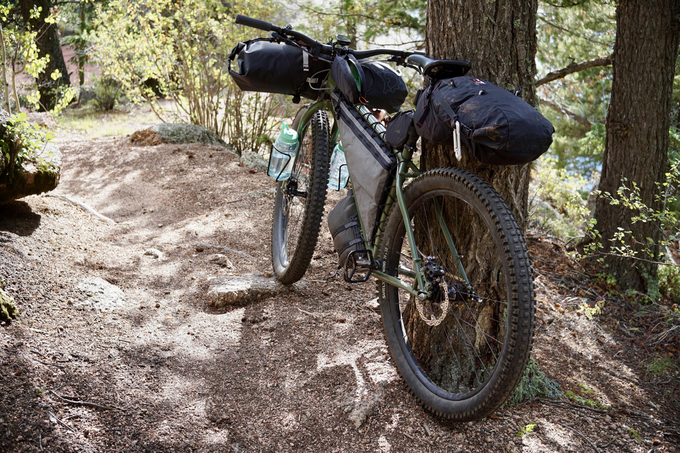 The loaded green Krampus leaned against a tree on singletrack, front three-quarter view showing the frame bag and handlebar roll