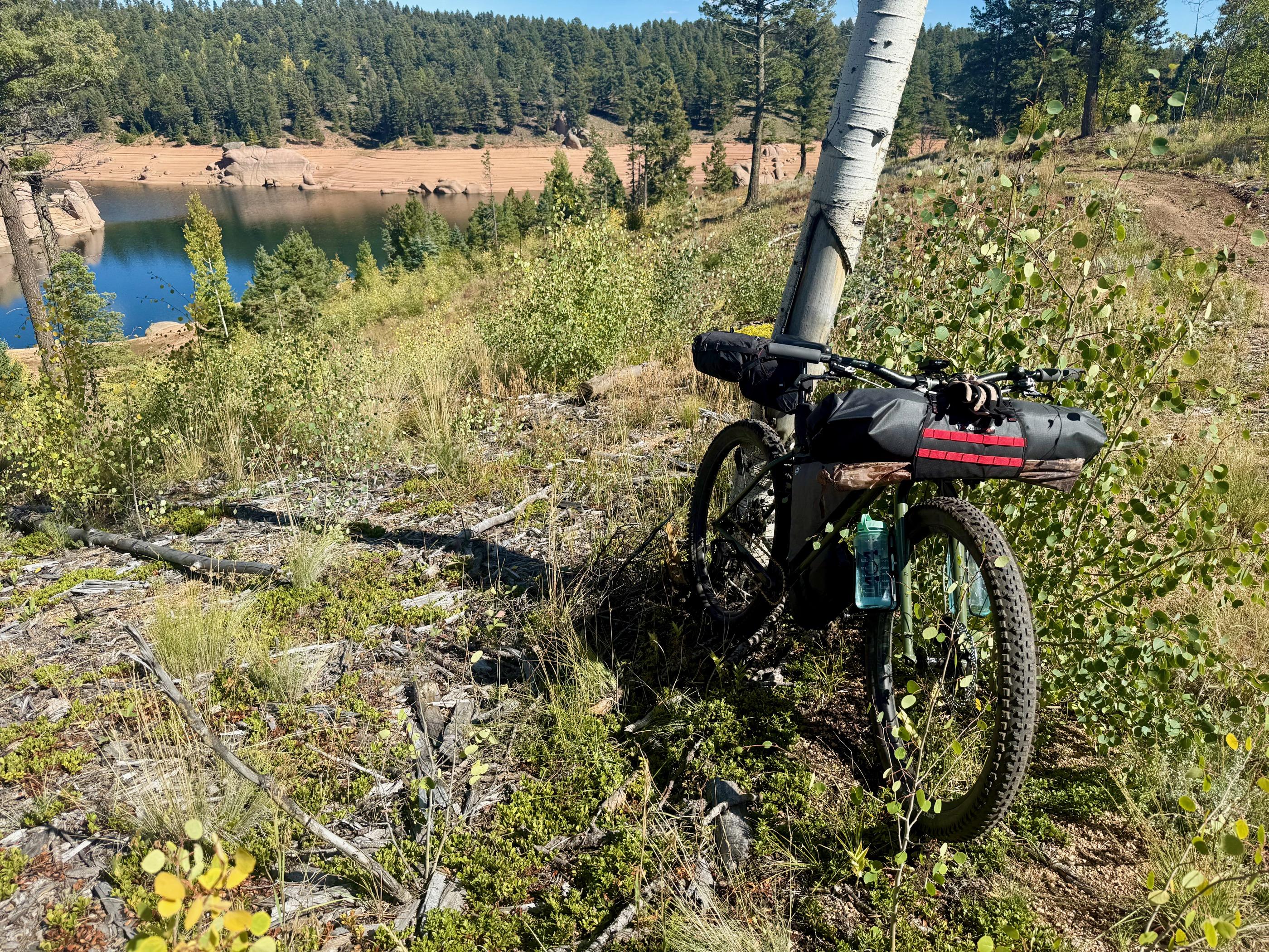 A loaded fat bike laid down on singletrack above Rampart Reservoir's drawn-down shoreline, aspen turning yellow on the far hillside