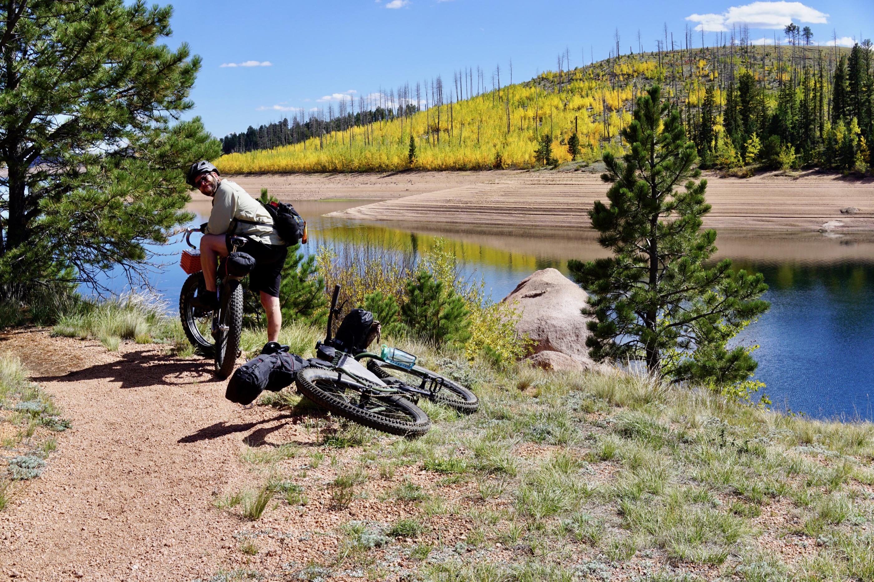 A rider pausing on a loaded fat bike above Rampart Reservoir's drawn-down shoreline, a second bike laid on the ground nearby, autumn hillside across the water