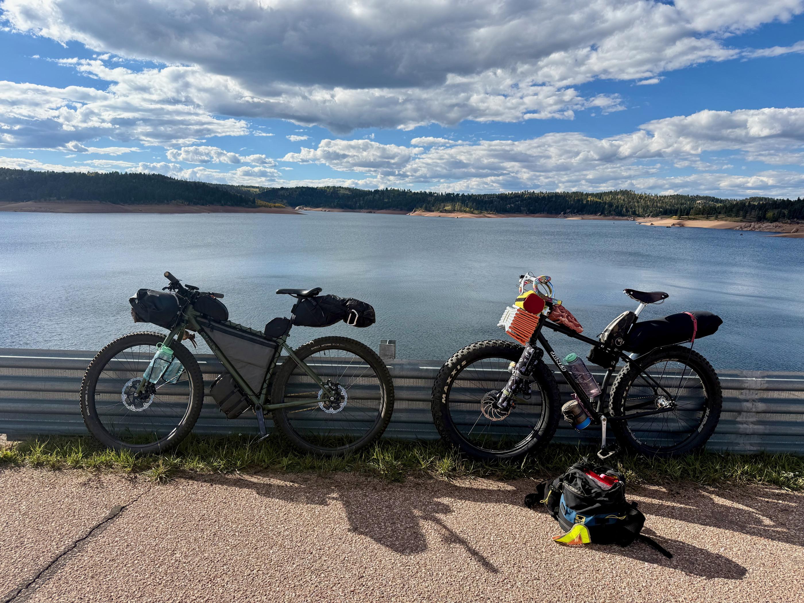 A loaded Surly Krampus and Fixie Dave's Jones fat bike leaned against a guardrail above Rampart Reservoir, water and forested hills behind them