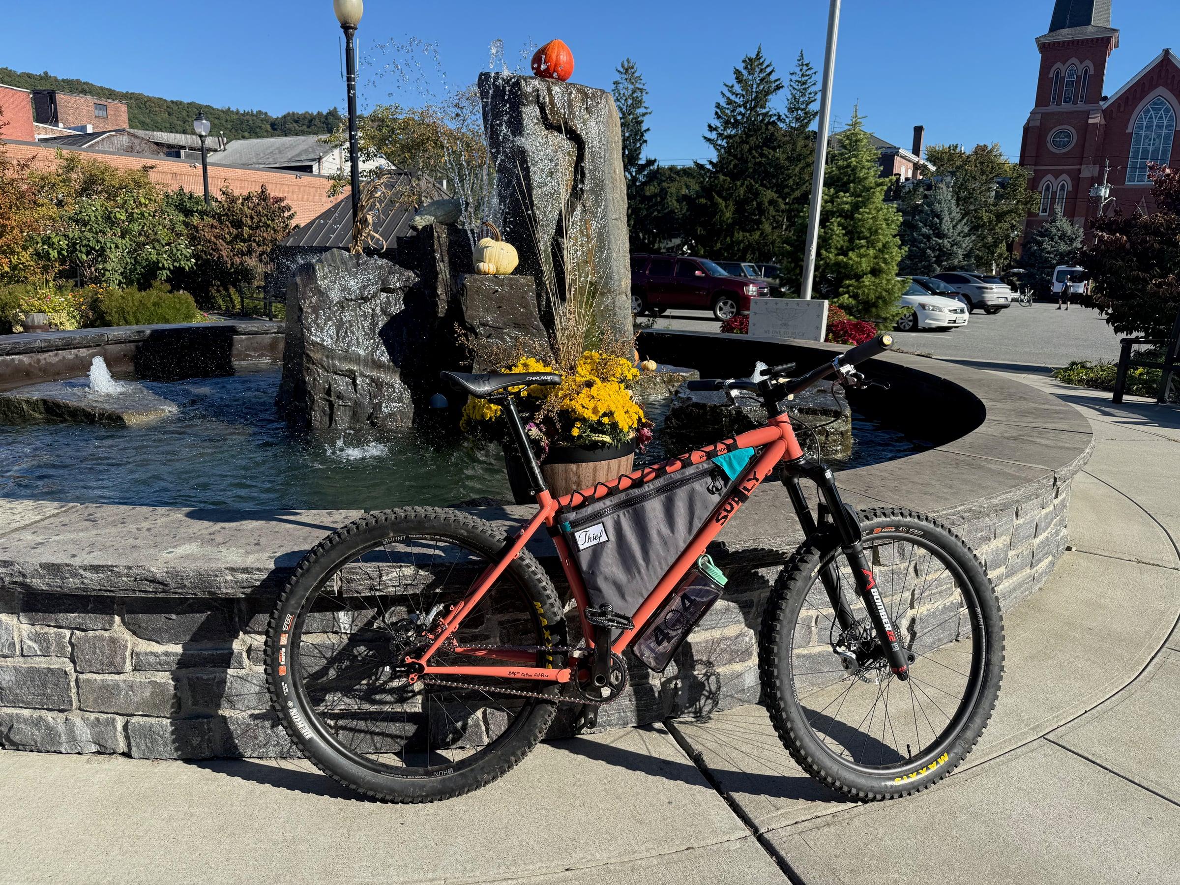 An orange Surly Karate Monkey with a frame bag and a suspension fork leaning against a stone fountain with pumpkins and yellow mums, with a brick church in the background