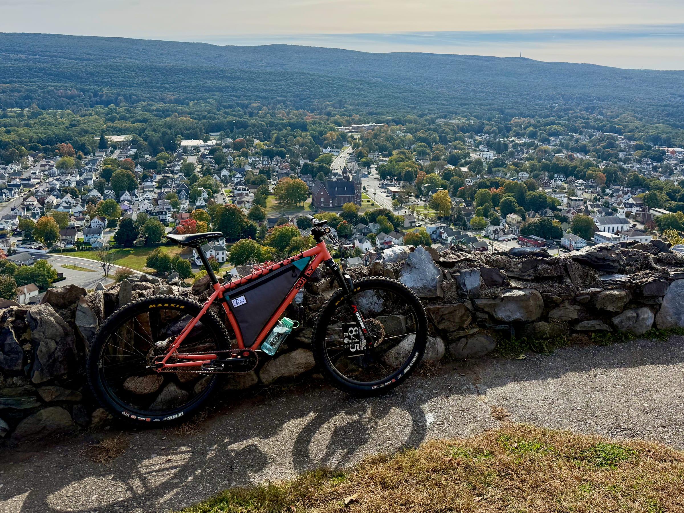 An orange Surly Karate Monkey with a race plate on the fork leaning against a stone wall at a lookout, with the rooftops of Port Jervis and a forested ridgeline spread out below