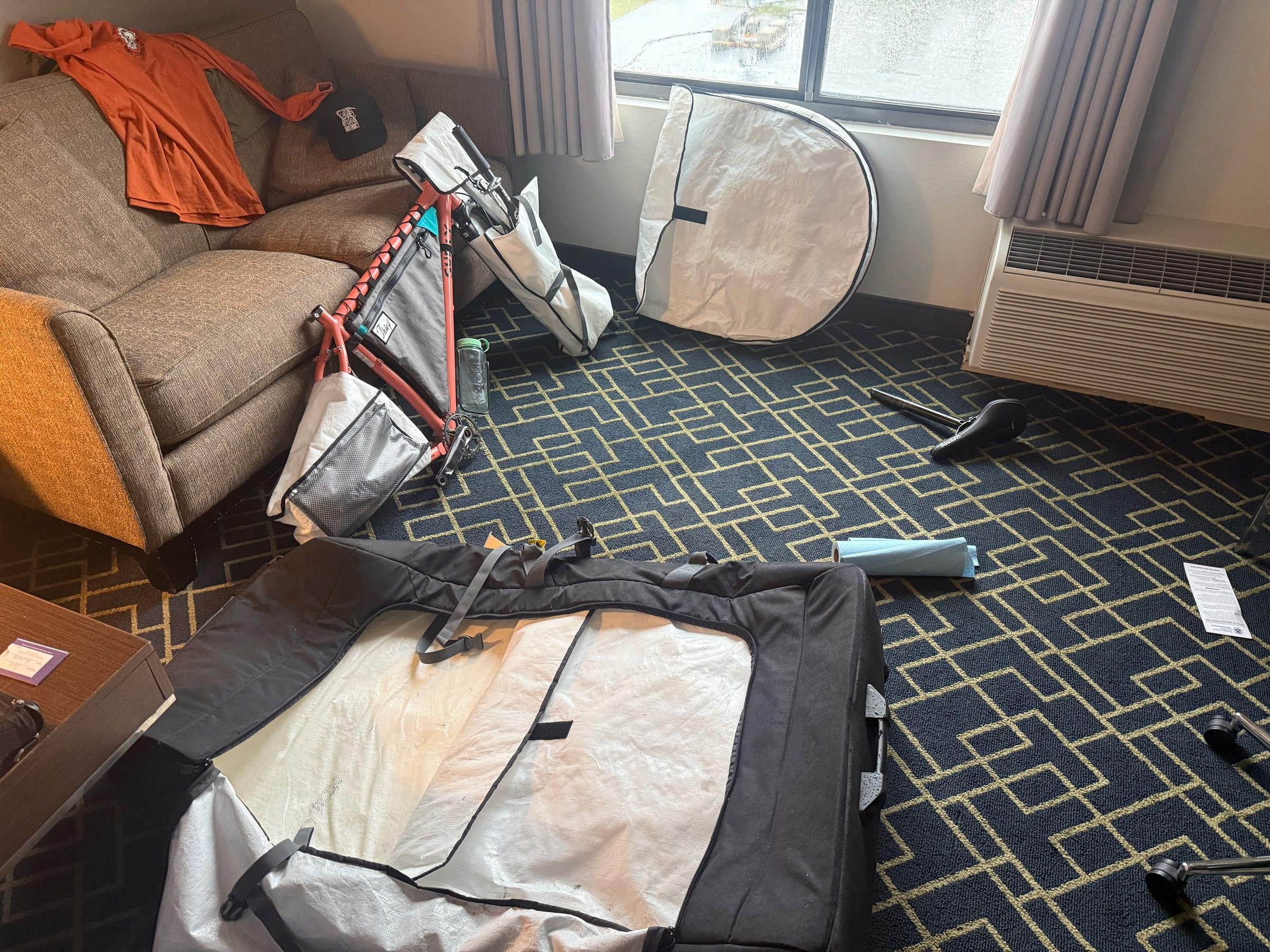 An orange Surly Karate Monkey frame leaning against a hotel couch next to wheel bags, with the saddle and seatpost lying on the carpet beside an open bike bag