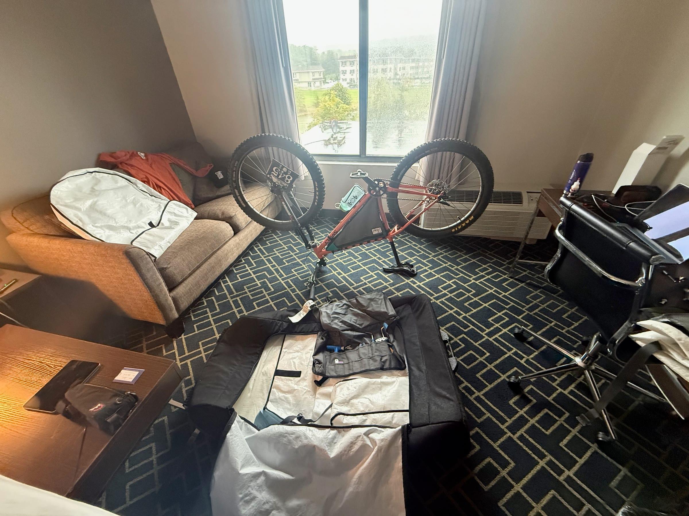 An orange Surly Karate Monkey upside down on its saddle and handlebars in a hotel room, with a race plate on the front wheel, an open bike bag and a tool roll on the floor, and rain on the window behind it